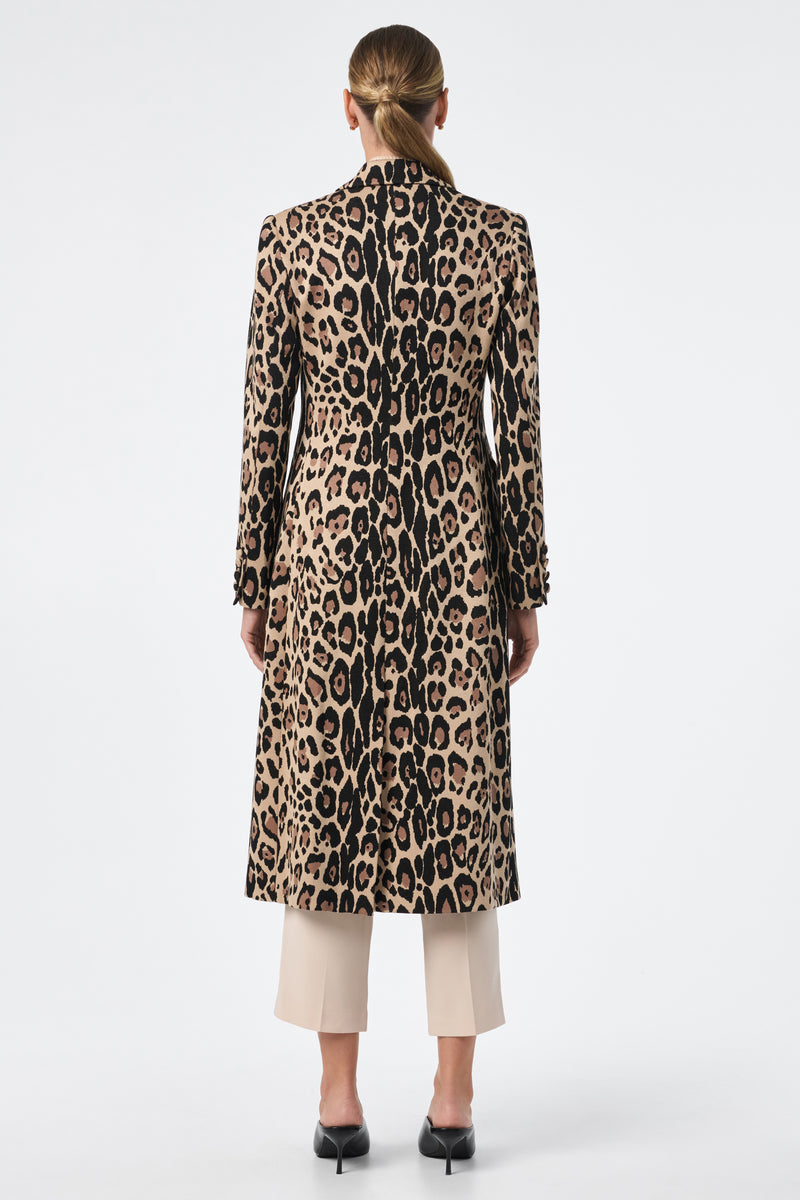 A person with long hair tied back is shown from behind, wearing the Playbill Coat—a long, fitted leopard print piece—over beige cropped pants and black low-heeled shoes, against a plain white background.