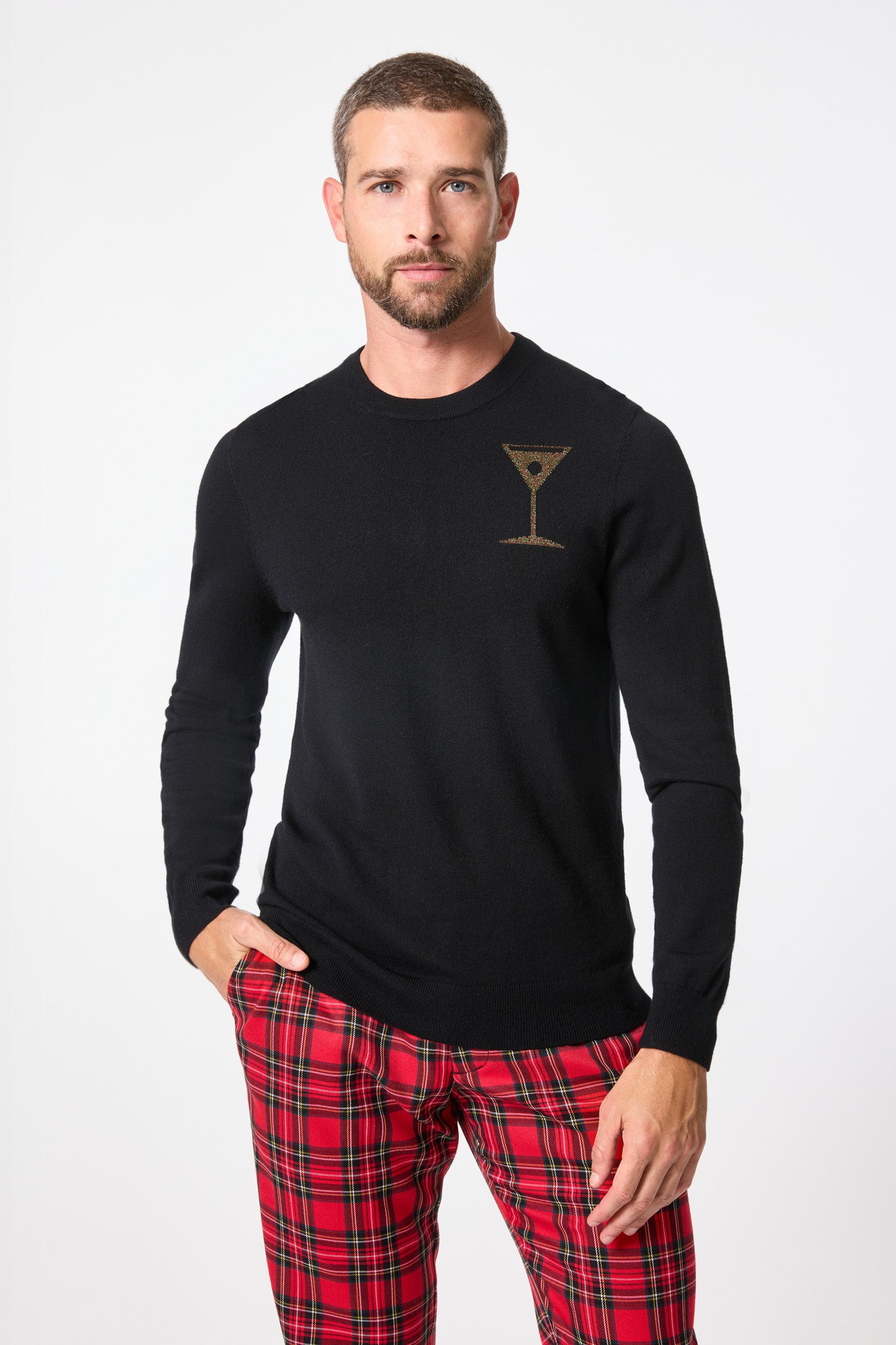 A man with short hair and a beard wears the black Martini Crewneck Sweater, featuring a martini glass graphic on the chest, paired with red plaid pants, standing against a plain white background.