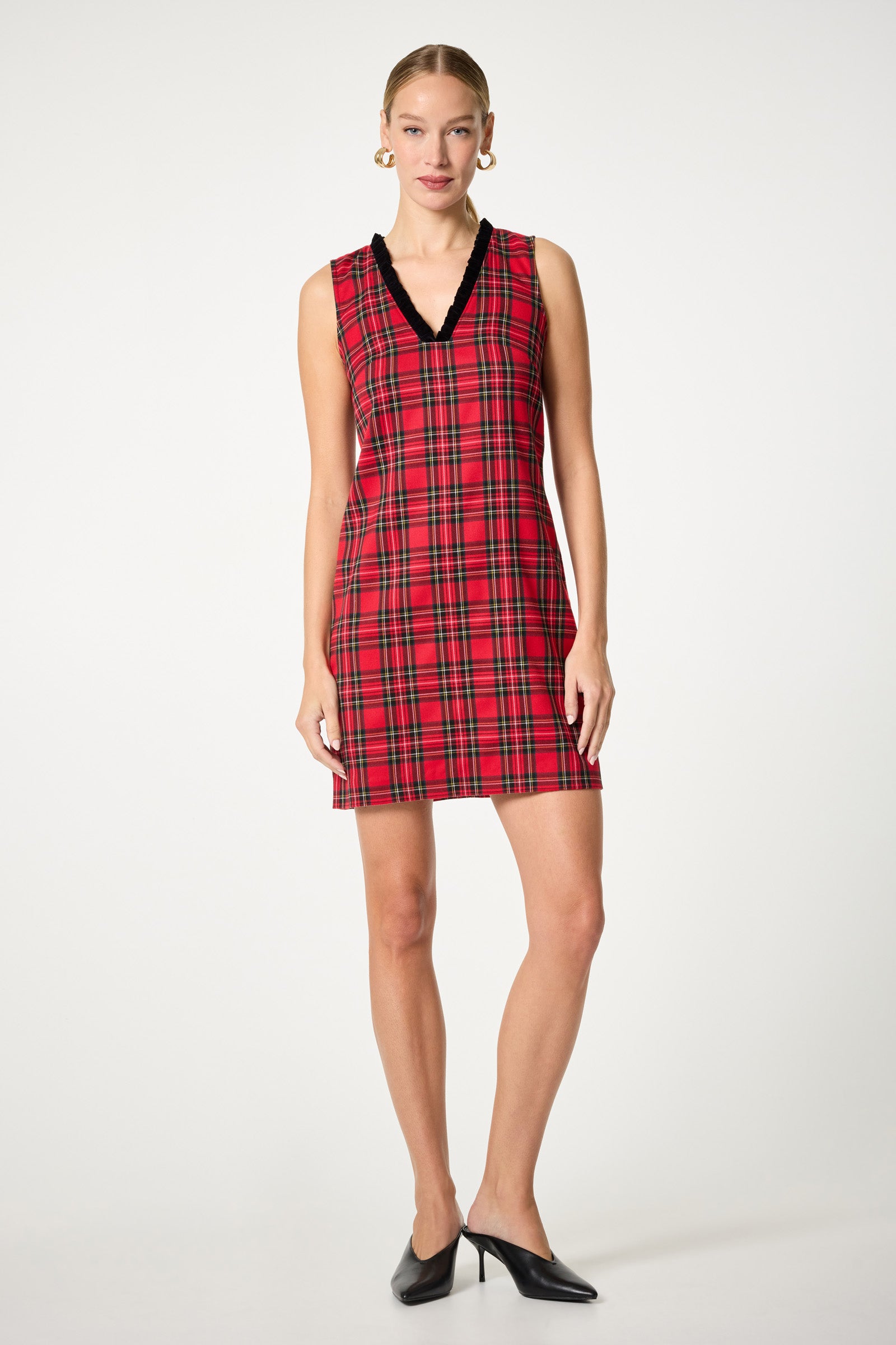 A woman stands against a plain background wearing the Revah Tartan Mini Dress, a sleeveless red plaid dress with black V-neck trim. She accessorizes with gold hoop earrings, slicked-back hair, and black pointed-toe heels.