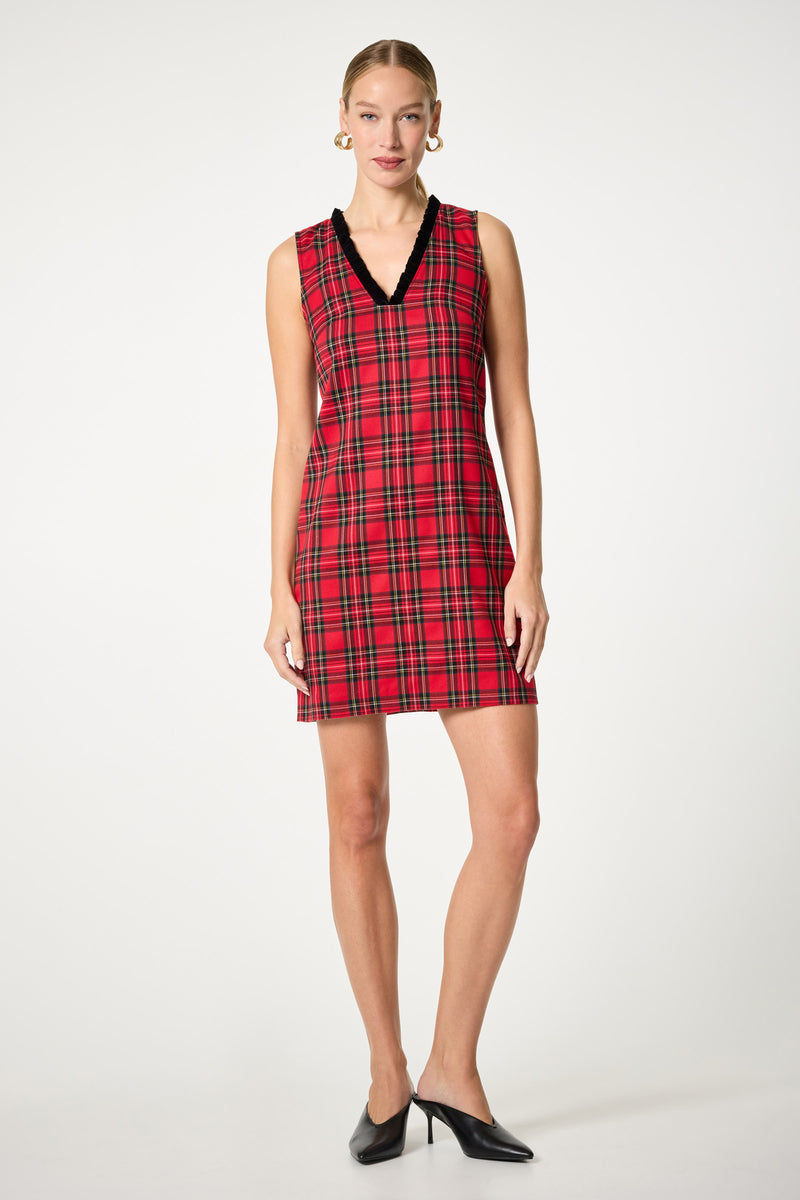 A woman stands against a plain background wearing the Revah Tartan Mini Dress, a sleeveless red plaid dress with black V-neck trim. She accessorizes with gold hoop earrings, slicked-back hair, and black pointed-toe heels.