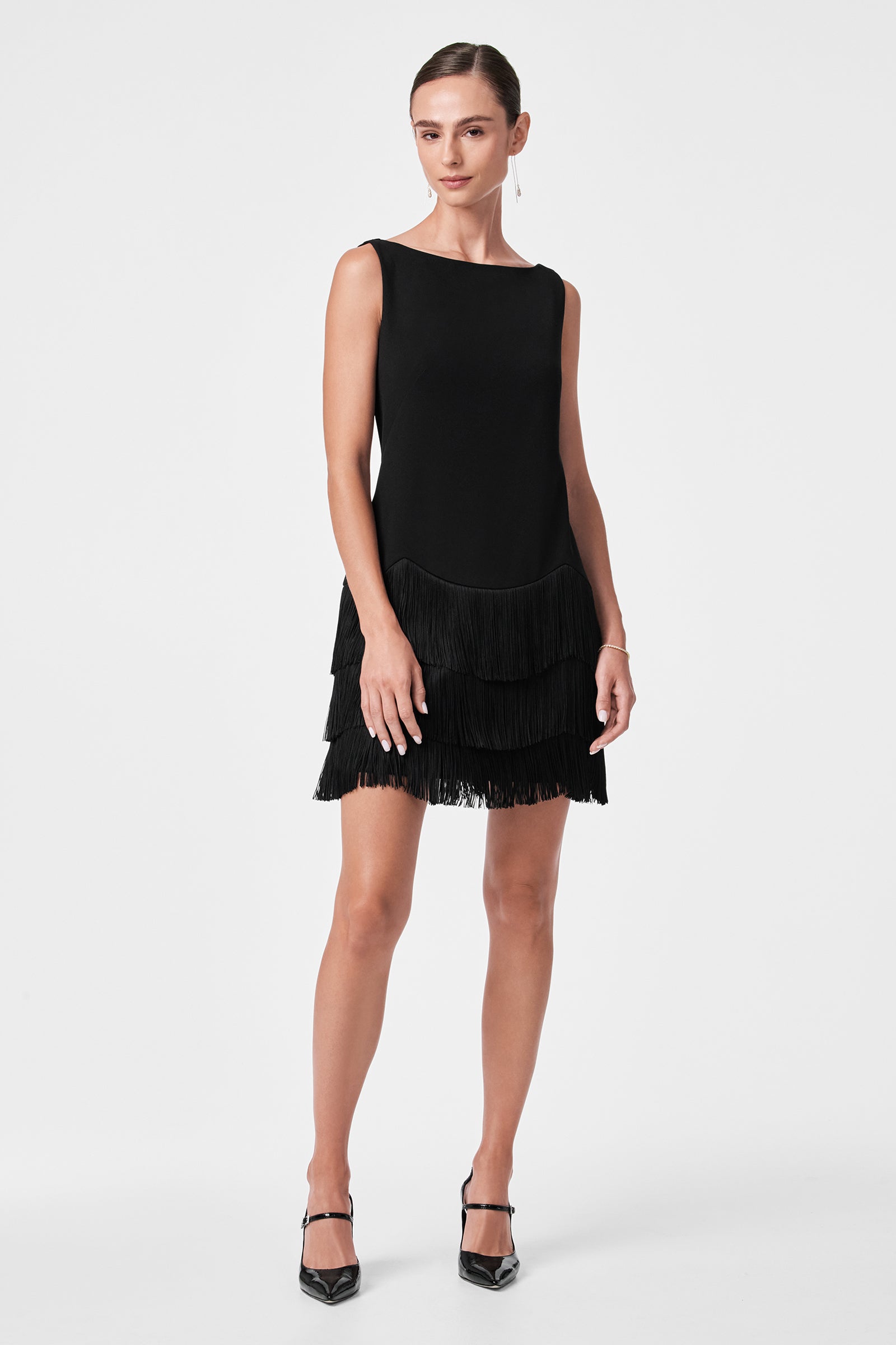 A woman stands against a white background wearing the sleeveless, knee-length Palais Dress with layered fringe details. Her hair is tied back, and she pairs the look with black pointed-toe heels.