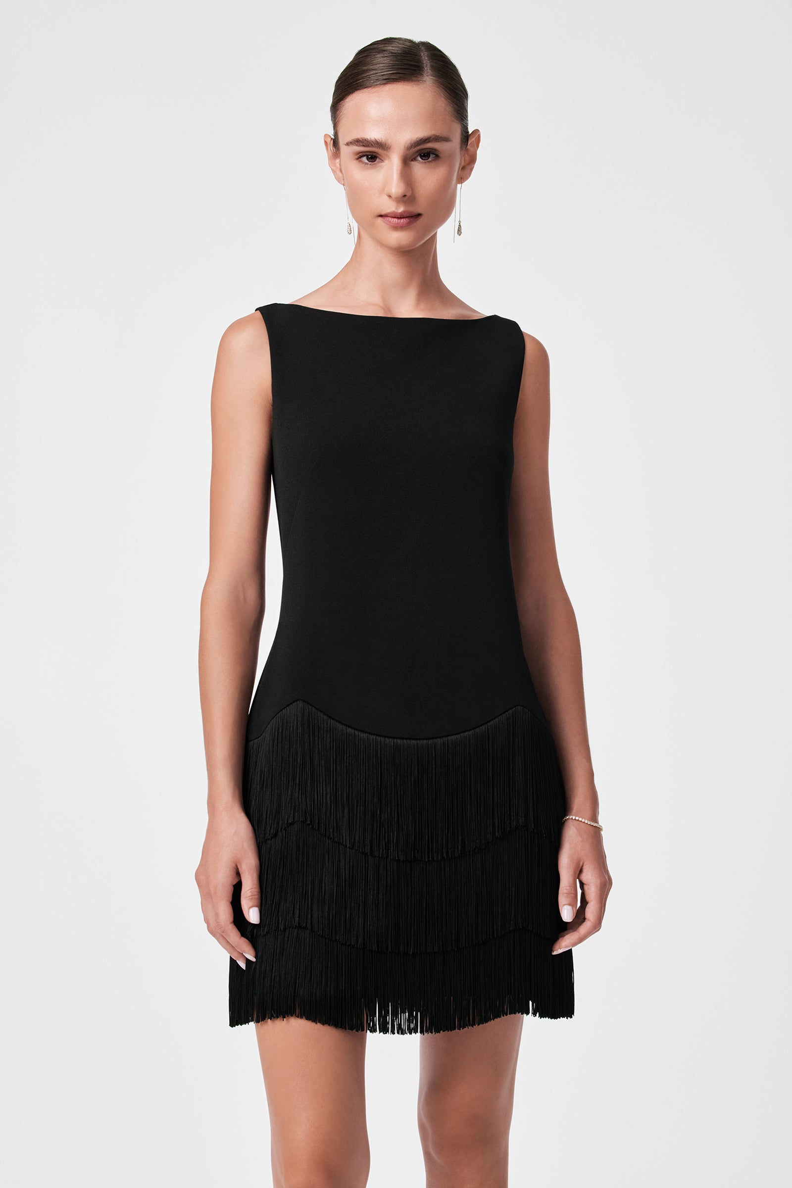 A woman models the Palais Dress, a sleeveless black dress featuring fringe details on the skirt. Her hair is neatly pulled back, and she accessorizes with subtle earrings and a bracelet against a plain white background.