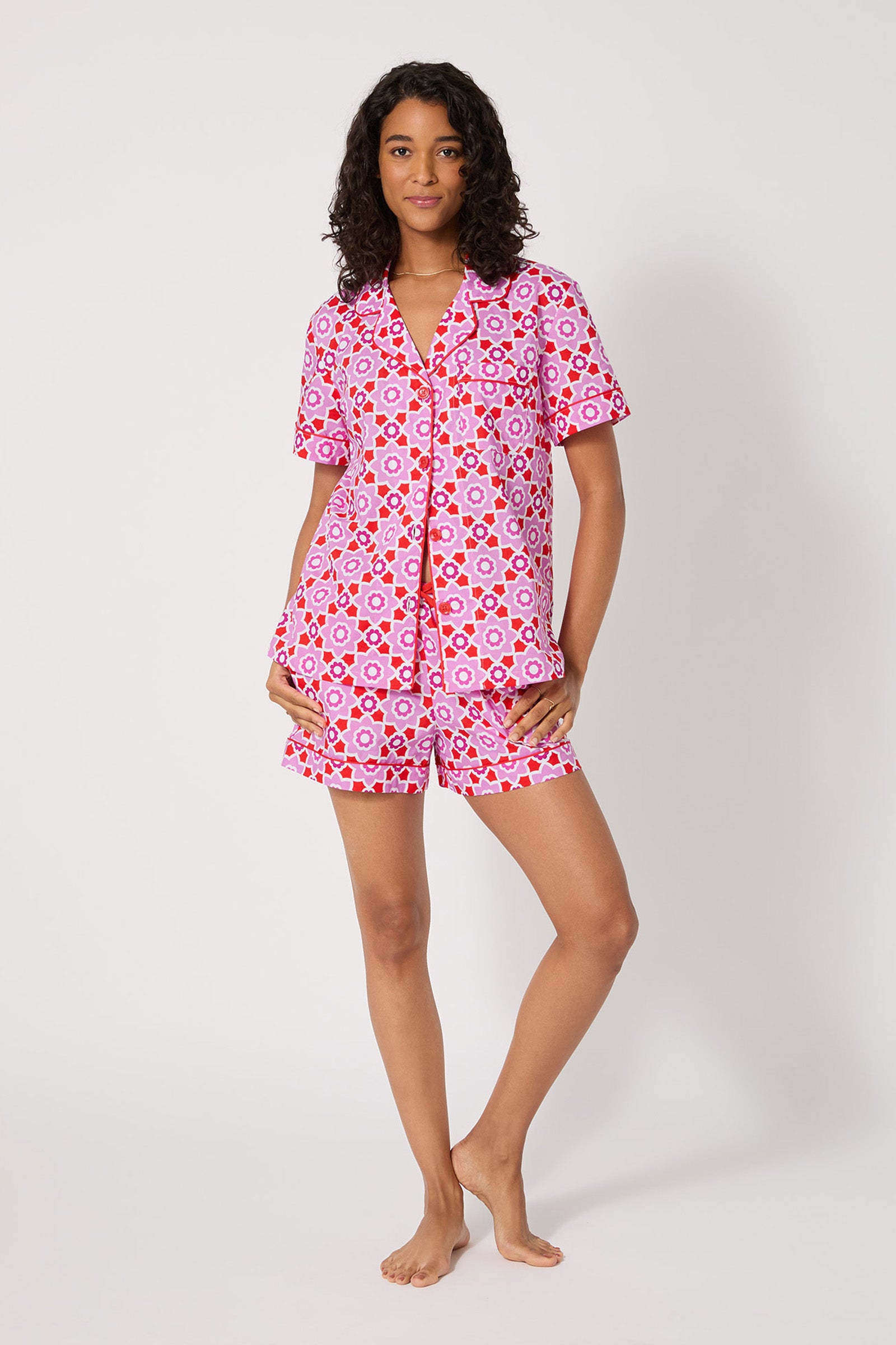 A woman stands barefoot against a white background, wearing the Shorty PJ Set - Mosaic Tile, a matching pink and red geometric-patterned short-sleeve pajama set with a button-up top and shorts.