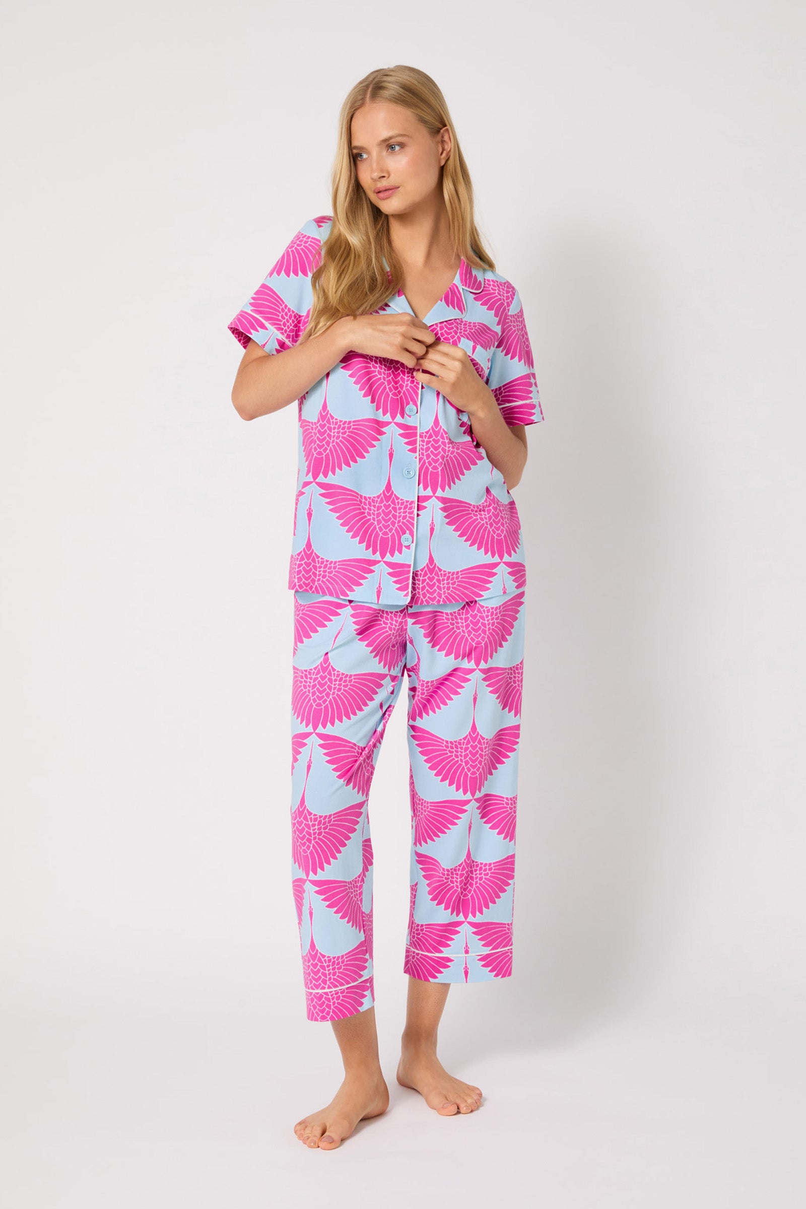 A woman with long blonde hair stands barefoot against a plain background, wearing the Crop PJ Set - Cranes: a matching short-sleeve and pants pajama set with a bright pink crane print on a light blue base, looking relaxed.