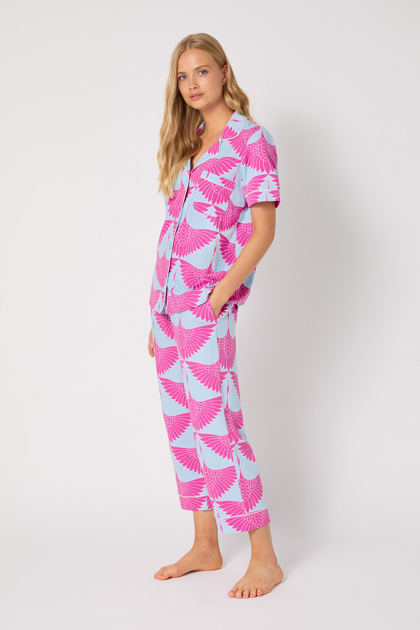 A woman stands barefoot against a white background, wearing the Crop PJ Set - Cranes, which features a matching light blue short-sleeve button-up top and loose pants with a bold pink crane-inspired pattern.