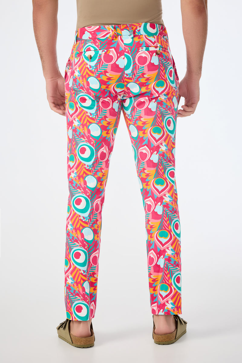 A person wearing the Clyde Slim Trouser - Dramont Herringbone, featuring bold peacock feather and abstract patterns in pink, green, orange, and white, stands against a plain white background, viewed from the back.