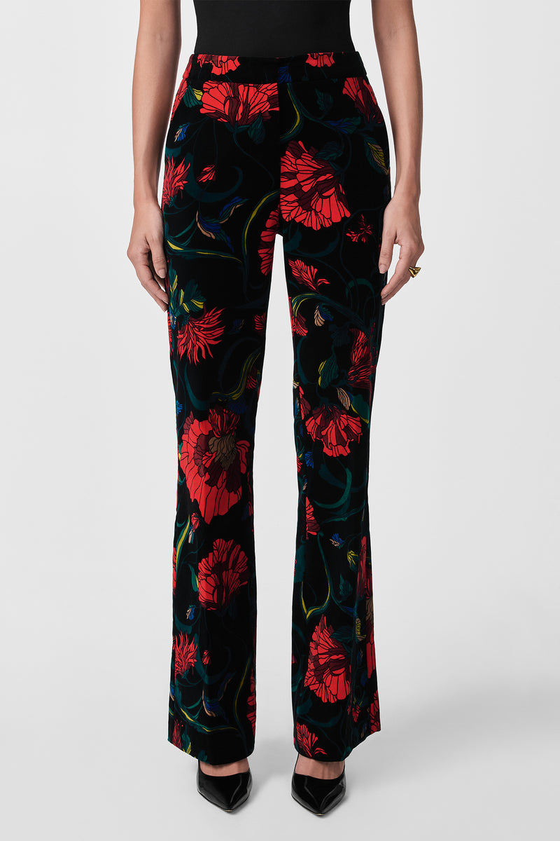A person wears Westport Printed Velvet Pants in black with a bold red floral print, standing against a plain white background with hands at their sides and paired with pointed black heels.