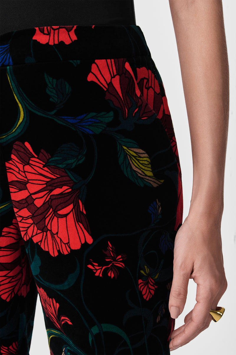 A close-up of a person wearing Westport Printed Velvet Pants featuring a bold red floral pattern, paired with a black top and gold ring. The background is plain and light-colored.