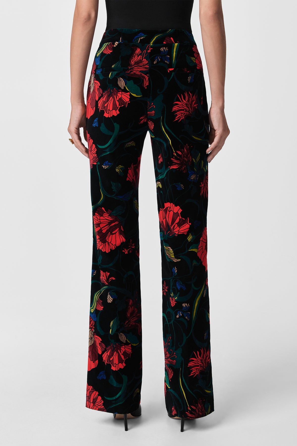 A person is seen from the back wearing Westport Printed Velvet Pants with a red and green floral design, styled with a black sleeveless top and black high-heeled shoes.