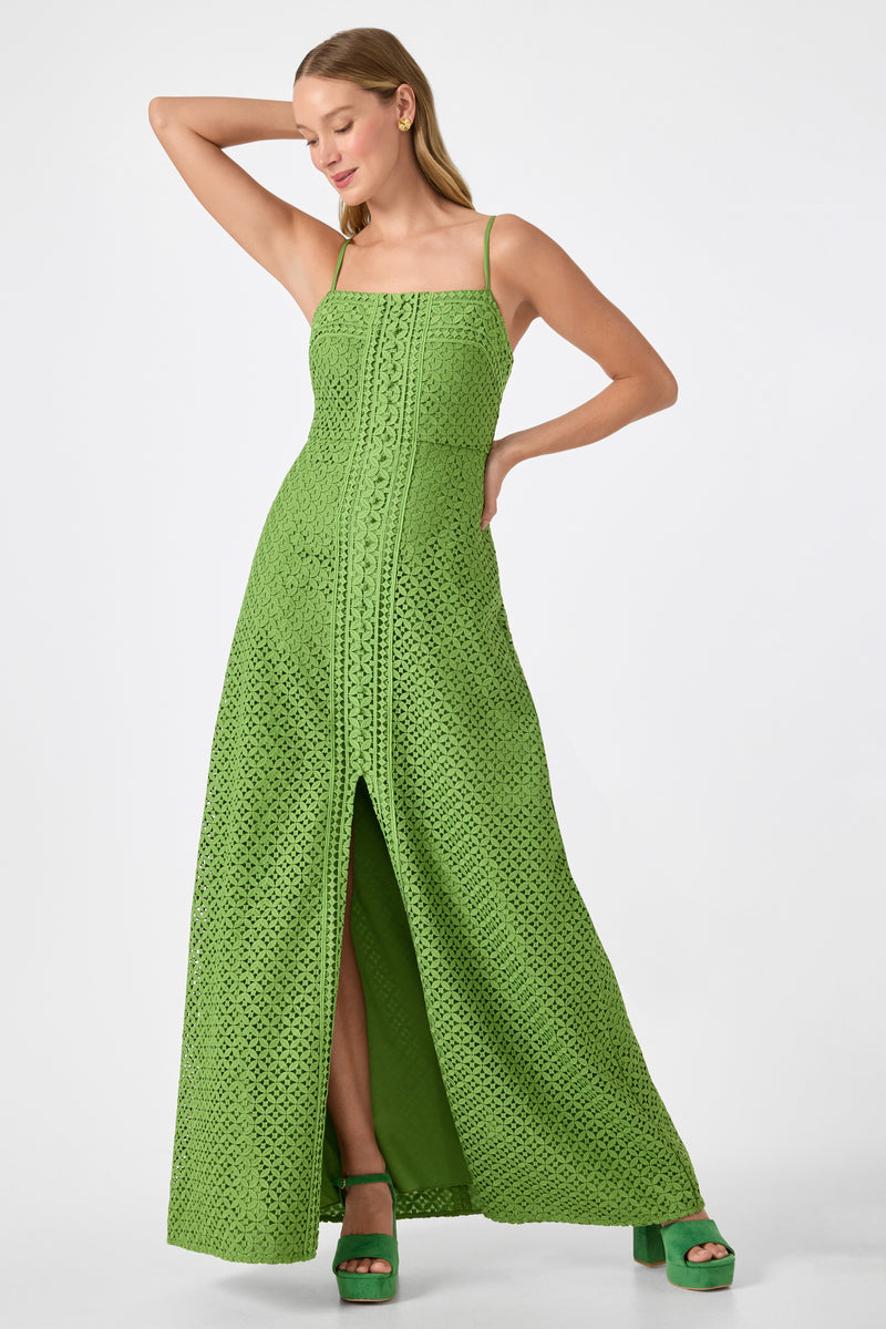 A woman poses in the Akumi Sleeveless Lace Maxi Dress, a long green crochet style with a front slit. She wears matching green platform heels, has one arm raised, head turned slightly, straight hair, and a subtle smile.