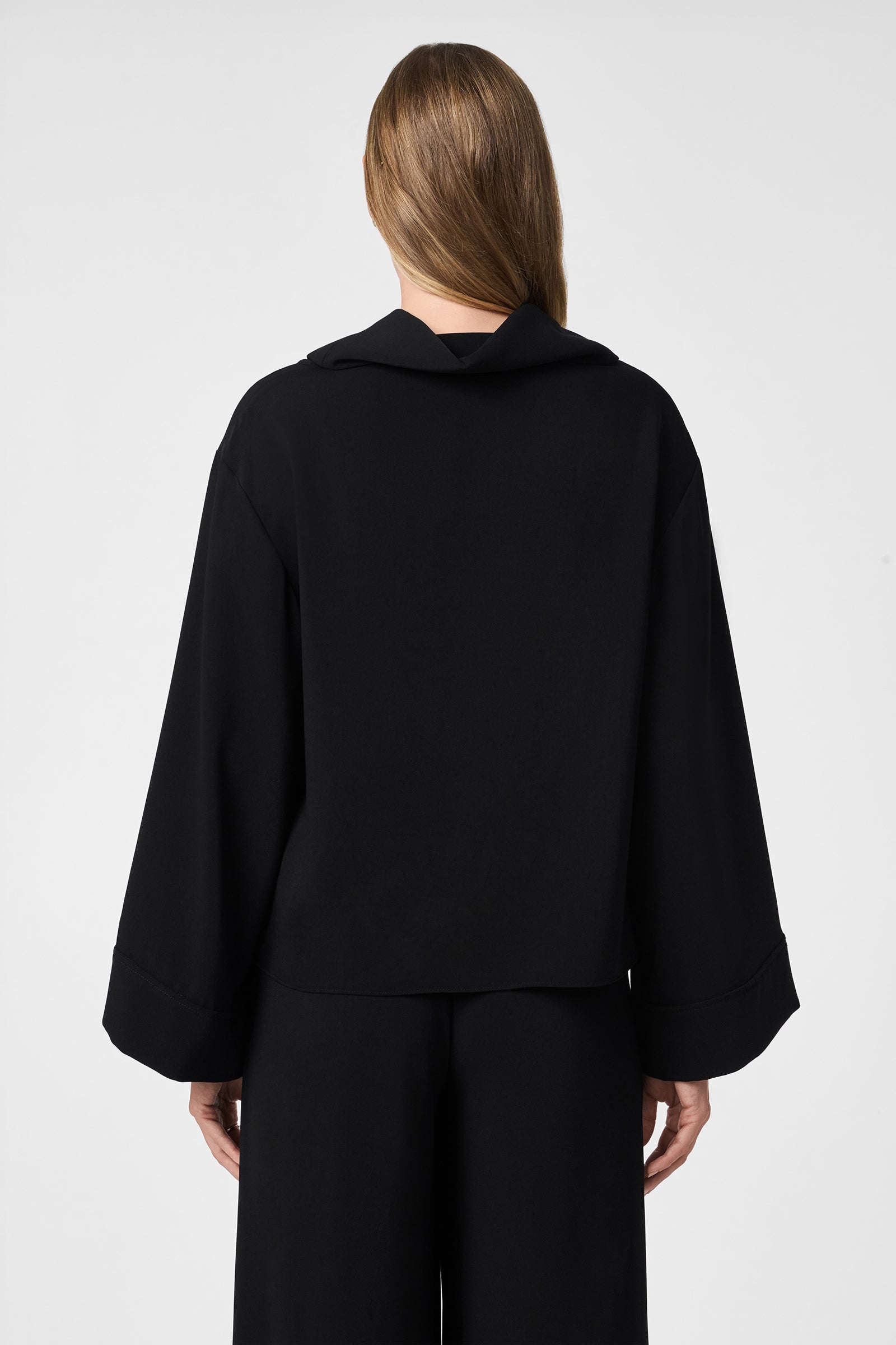 A person with long, light brown hair is seen from behind wearing the Staged Top—a loose-fitting black top with wide sleeves—paired with matching black pants, set against a plain light background.