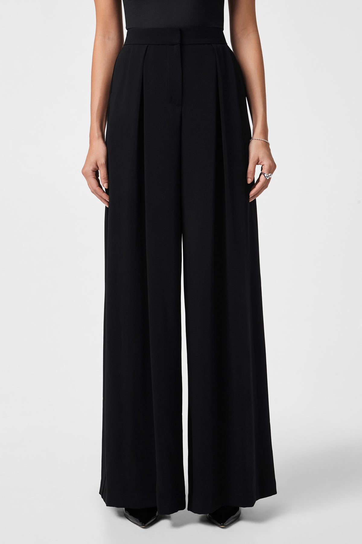 A person models the Saige Long Pant, a high-waisted, wide-leg black dress pant, paired with a sleeveless black top. Only their lower torso, legs, and hands are shown against a plain white background.