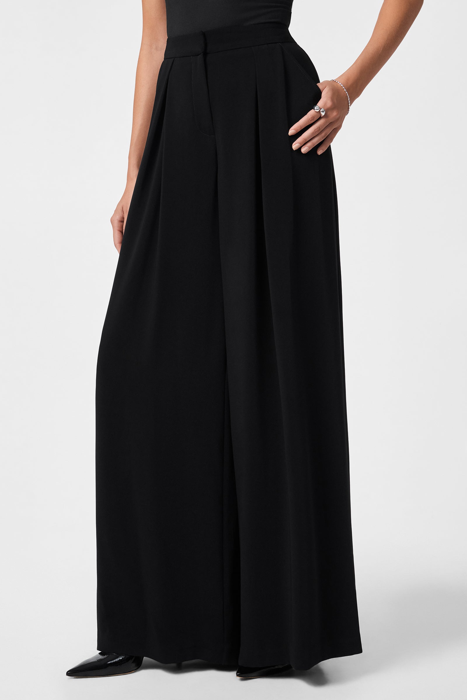 Someone wearing the Saige Long Pant—a high-waisted, wide-leg black pant—paired with a black top, black pointed shoes, and a bracelet, stands against a plain light background.