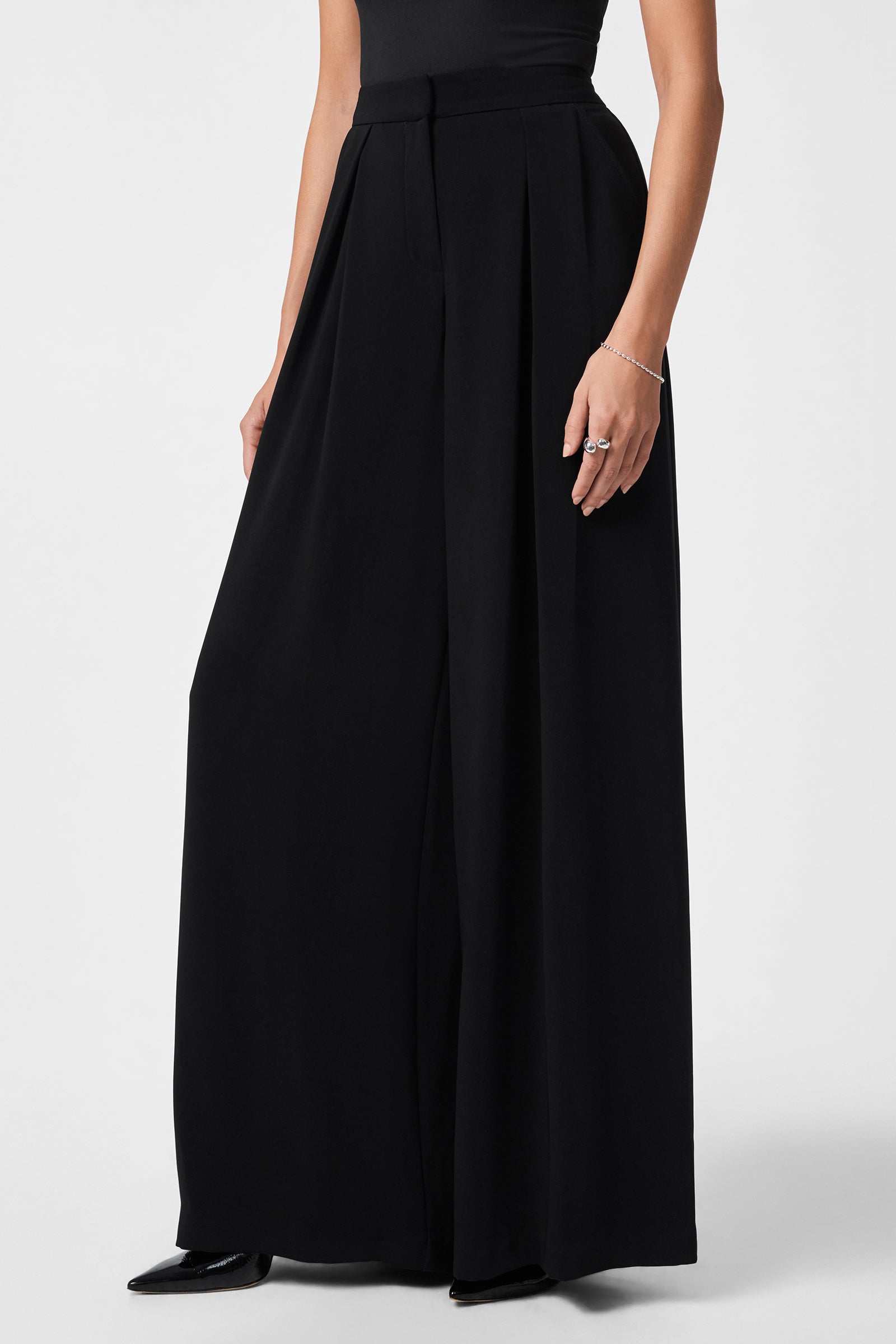 A person models the Saige Pant—high-waisted, black wide-leg trousers—with a fitted black sleeveless top, black pointed-toe shoes, and silver jewelry, set against a plain light background.