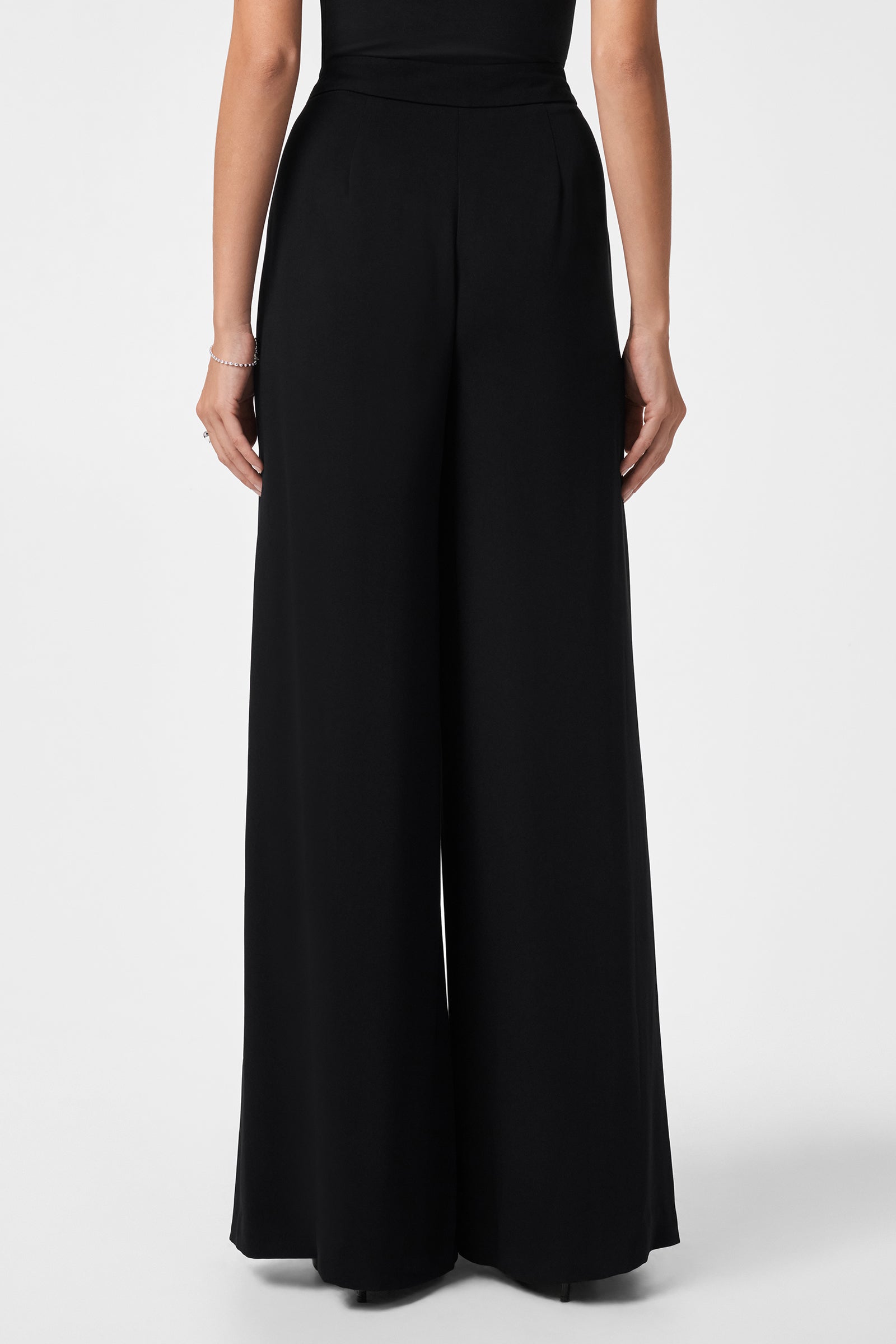 A person wearing the Saige Long Pant—high-waisted, wide-leg black pants—and a black top is shown from the back against a plain white background.