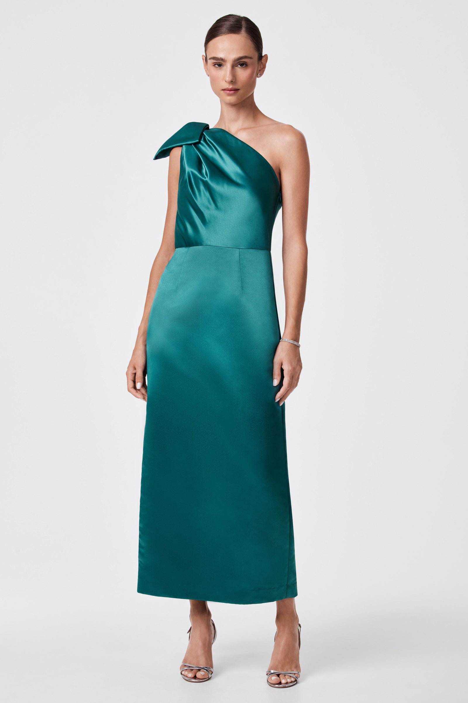A woman models the Romance Dress—a teal, one-shoulder satin gown with a dramatic shoulder bow, fitted bodice, and straight ankle-length skirt. She pairs it with silver heels and minimal jewelry against a simple backdrop.