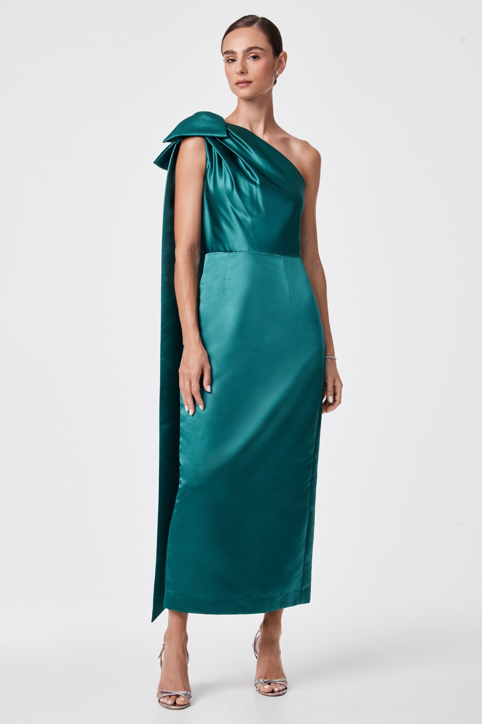 A woman stands against a white background wearing the teal, one-shoulder satin Romance Dress with a large bow on the shoulder. Her hair is tied back, and she pairs the look with silver open-toe high-heeled sandals.