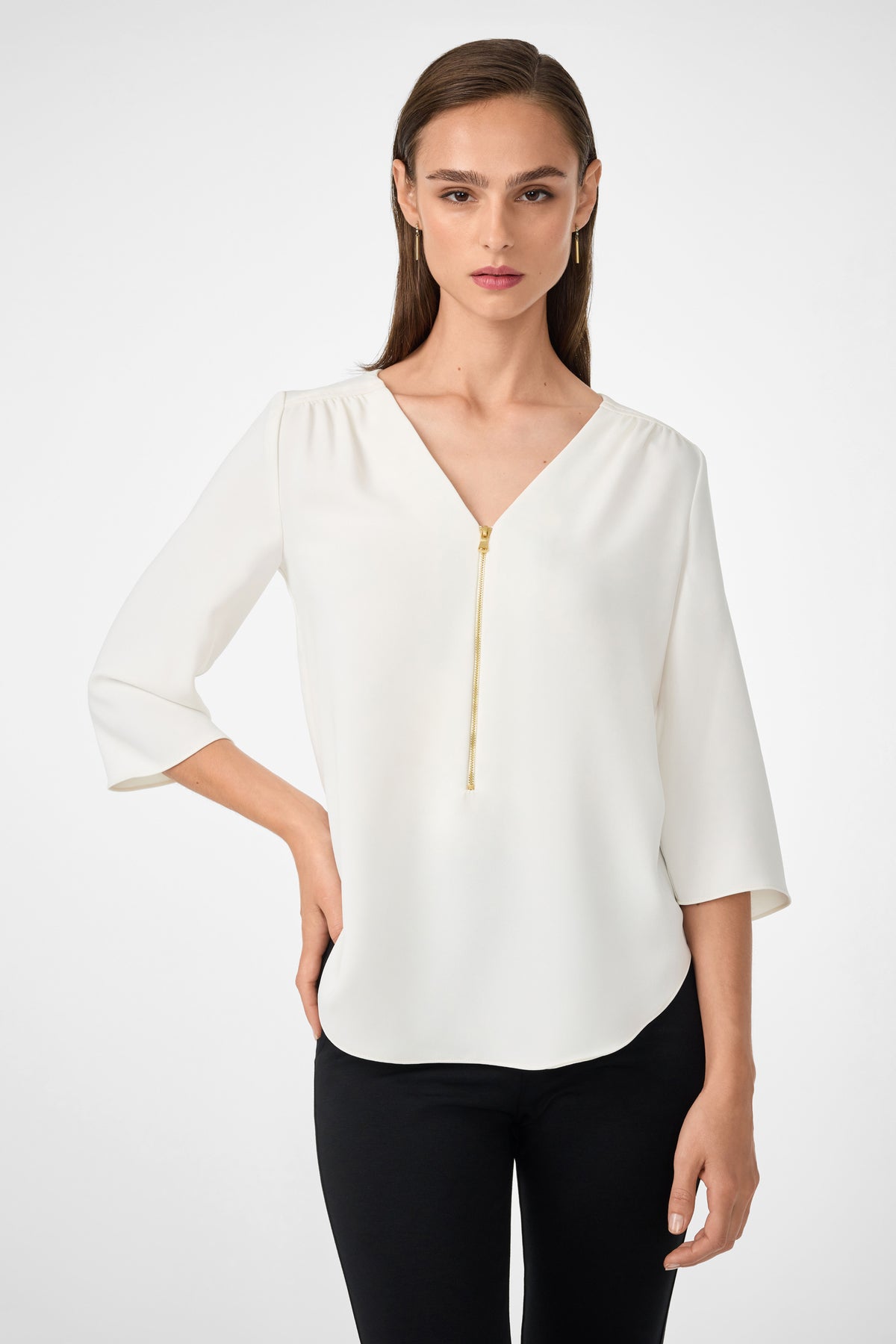 A woman wears the Tamarac Top in Whitewash—a white, long-sleeve blouse with a V-neckline and gold zipper detail—paired with black pants against a plain light background.