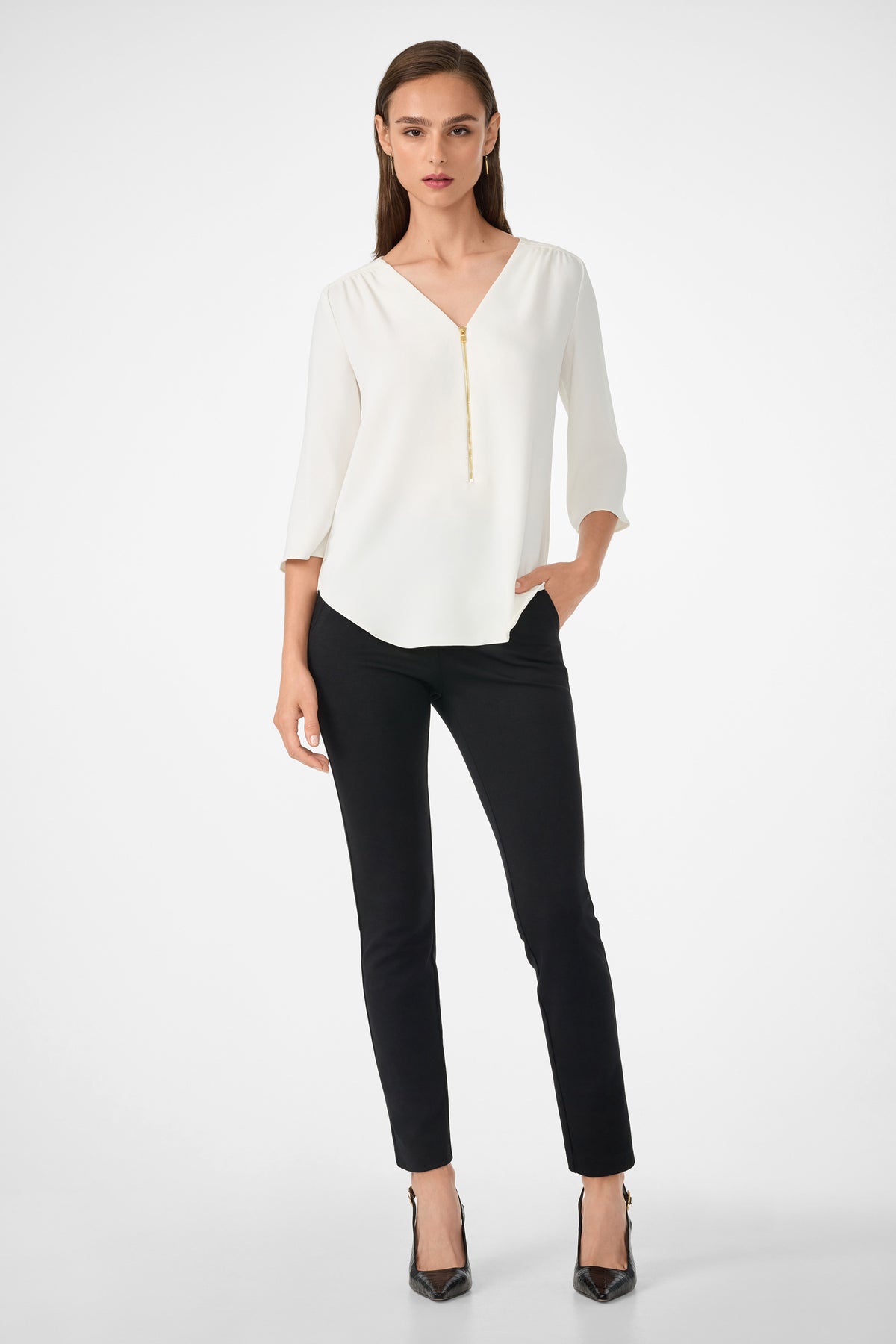 A woman stands against a plain background, wearing the Tamarac Top—a white V-neck blouse with a front zipper—paired with black slim-fit pants and heels. One hand is in her pocket as she looks confidently at the camera.