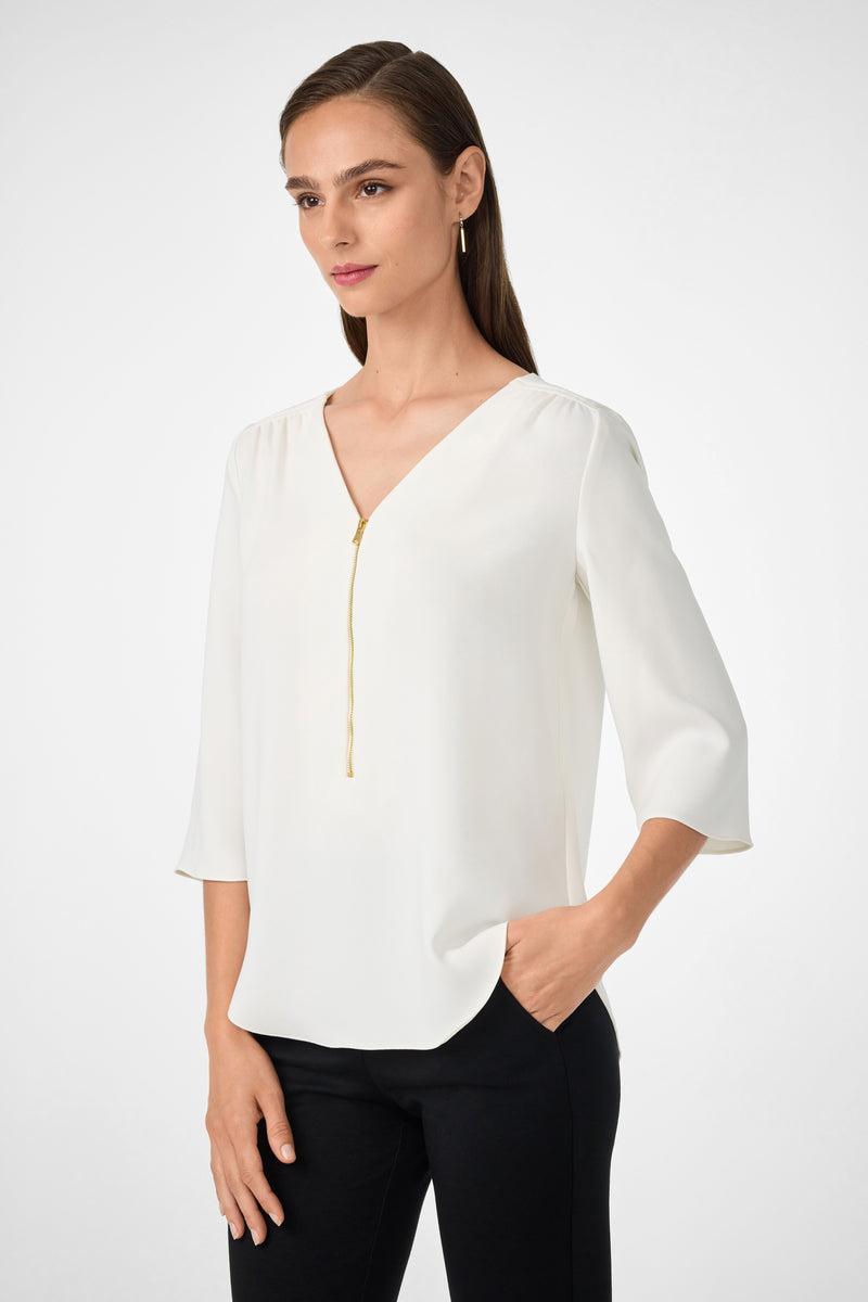 A woman with straight brown hair, tucked behind her ear, stands against a plain light background wearing the Tamarac Top—a white V-neck blouse with three-quarter sleeves—and black pants, with her left hand in her pocket.