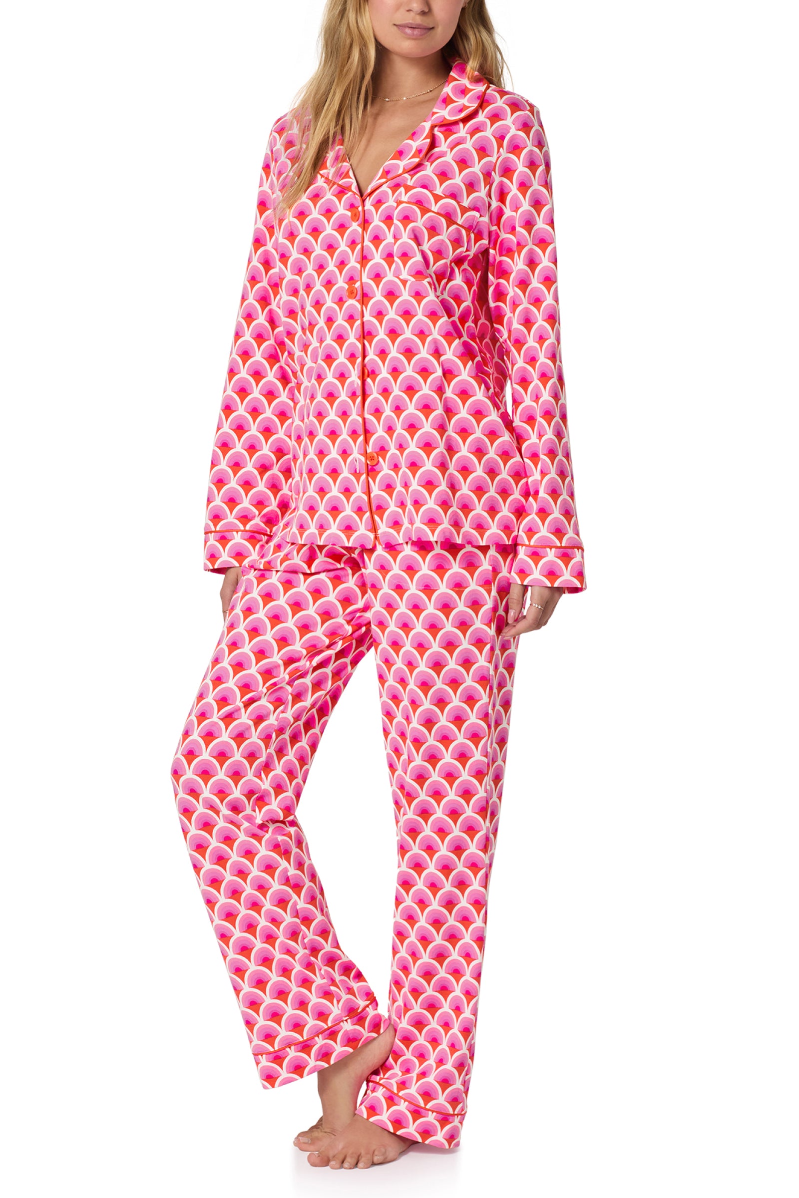 A woman stands barefoot against a white background wearing the Classic PJ Set - Scallop, a matching long-sleeve button-up pajama set featuring bright pink, orange, and white scallop patterns.