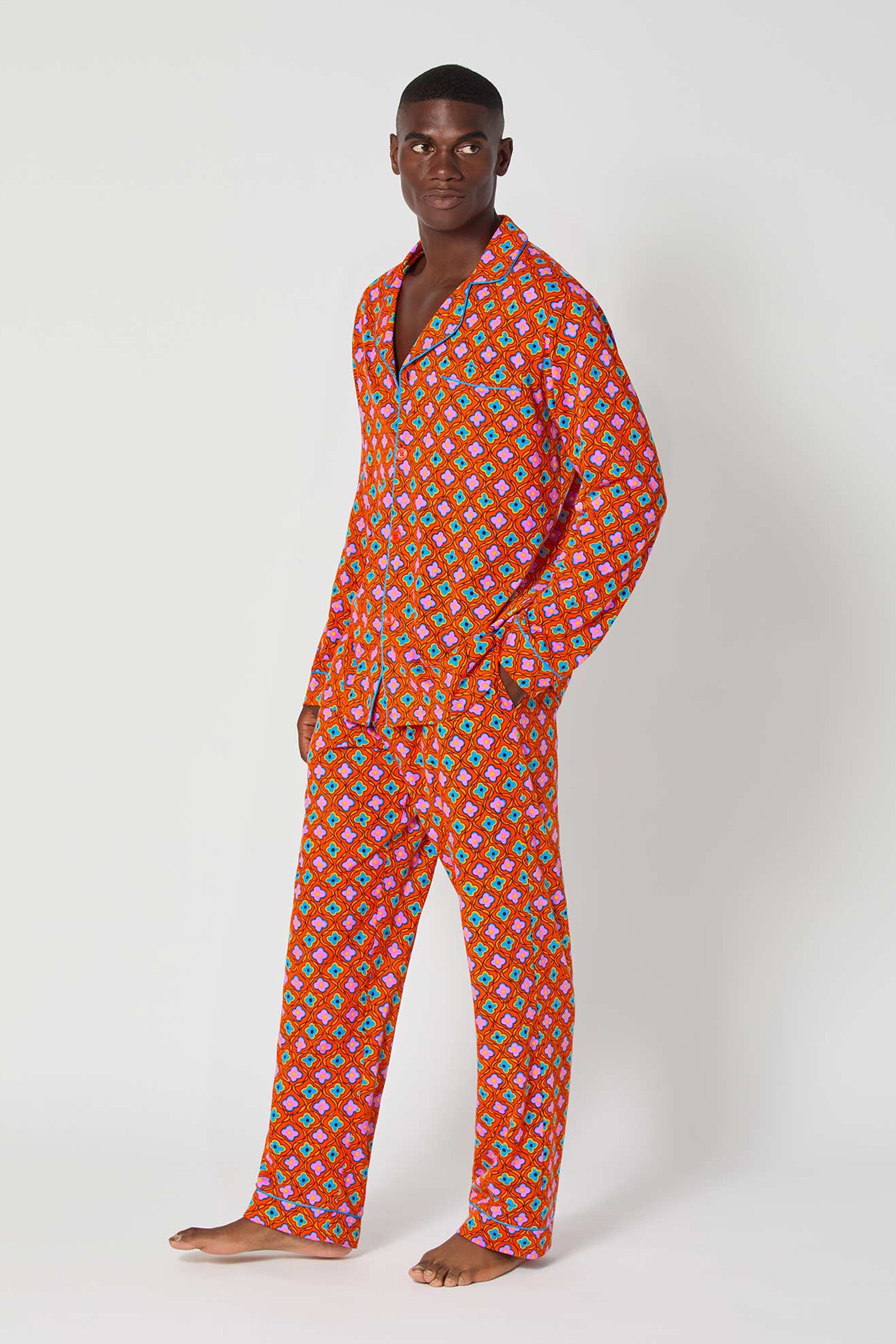 A man stands barefoot against a white background, wearing the Daisy Foulard Mens Classic Pj—a matching red pajama set with a colorful geometric pattern. He looks slightly to the side with a relaxed expression.