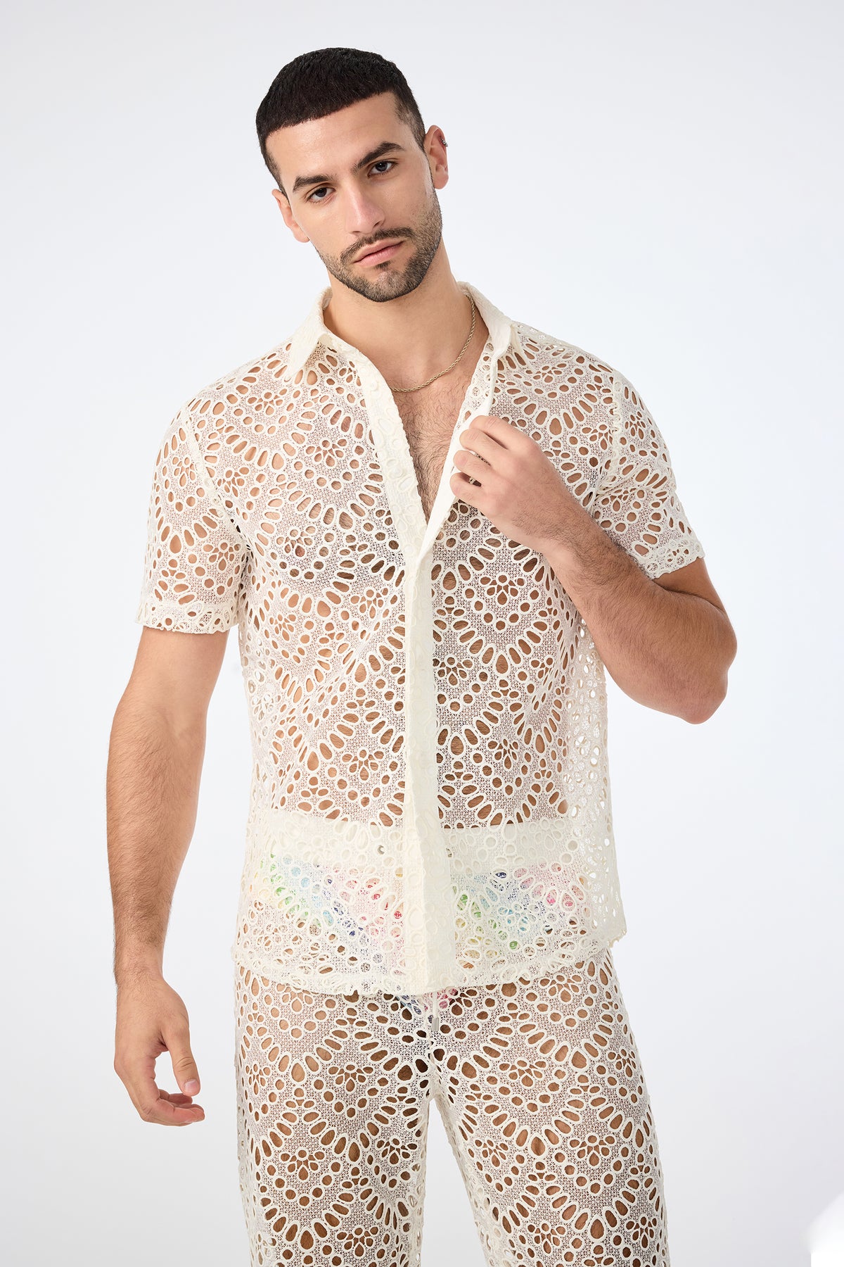 A man with short dark hair and a trimmed beard poses against a plain background, wearing the Long Beach Short-Sleeve Shirt in Whitewash with matching lace-patterned pants that reveal colorful underwear beneath.