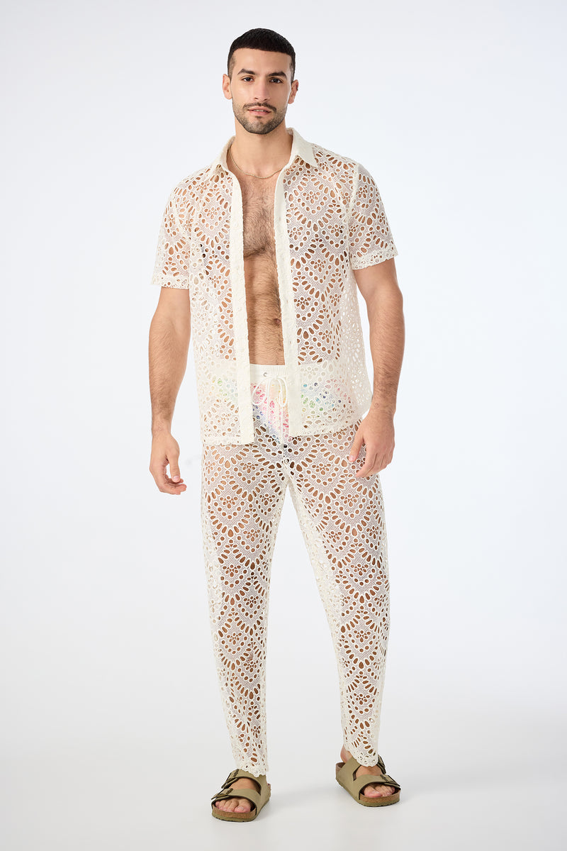 A man stands against a plain background wearing the Venice Beach Resort Pant in Lerins Lace, paired with a matching sheer white lace short-sleeve shirt over pastel swim trunks and green sandals.