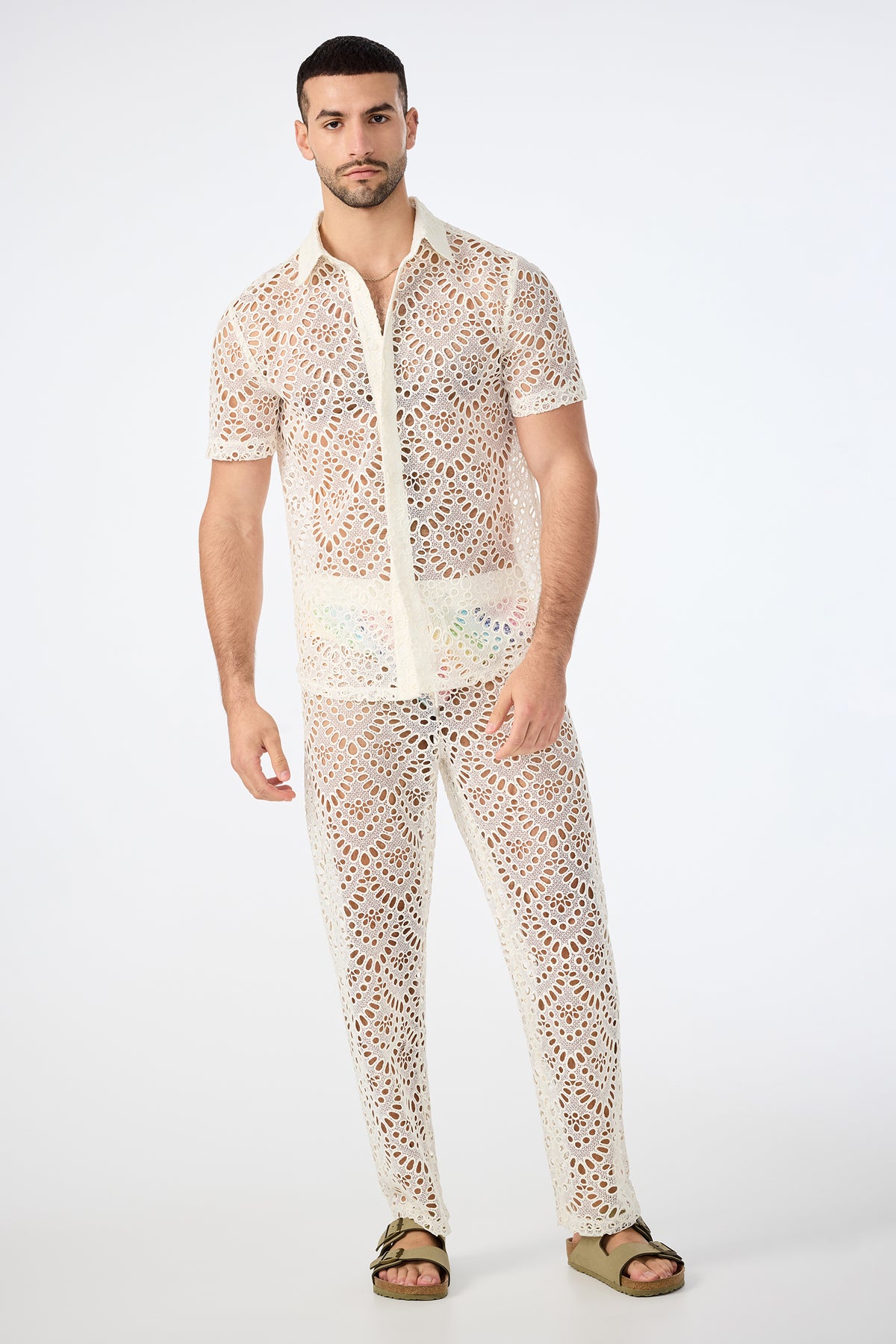 A man stands against a plain background wearing the Long Beach Short-Sleeve Shirt and matching white, see-through lace pants with a geometric pattern, paired with beige sandals.