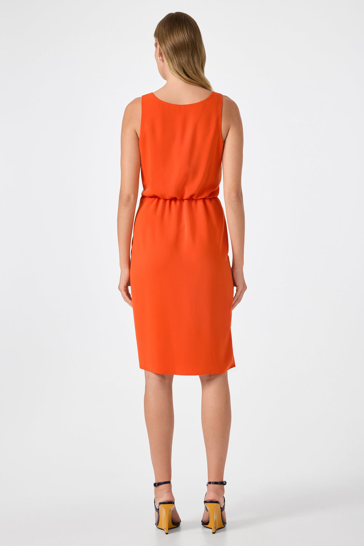 A woman with straight, light brown hair stands facing away in the ORita Sleeveless Tie-Waist Midi Dress and black high-heeled sandals against a plain white background.