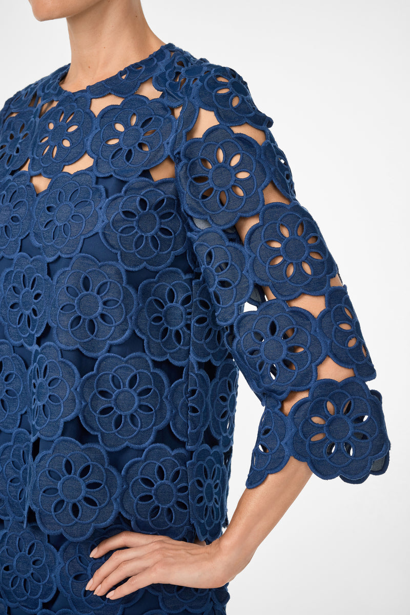 A person wears the Kabosu Top - Kusho Embroidery, featuring a navy blue color, large textured floral cut-out patterns, three-quarter sleeves, and a round neckline. Their hand is resting on their hip.