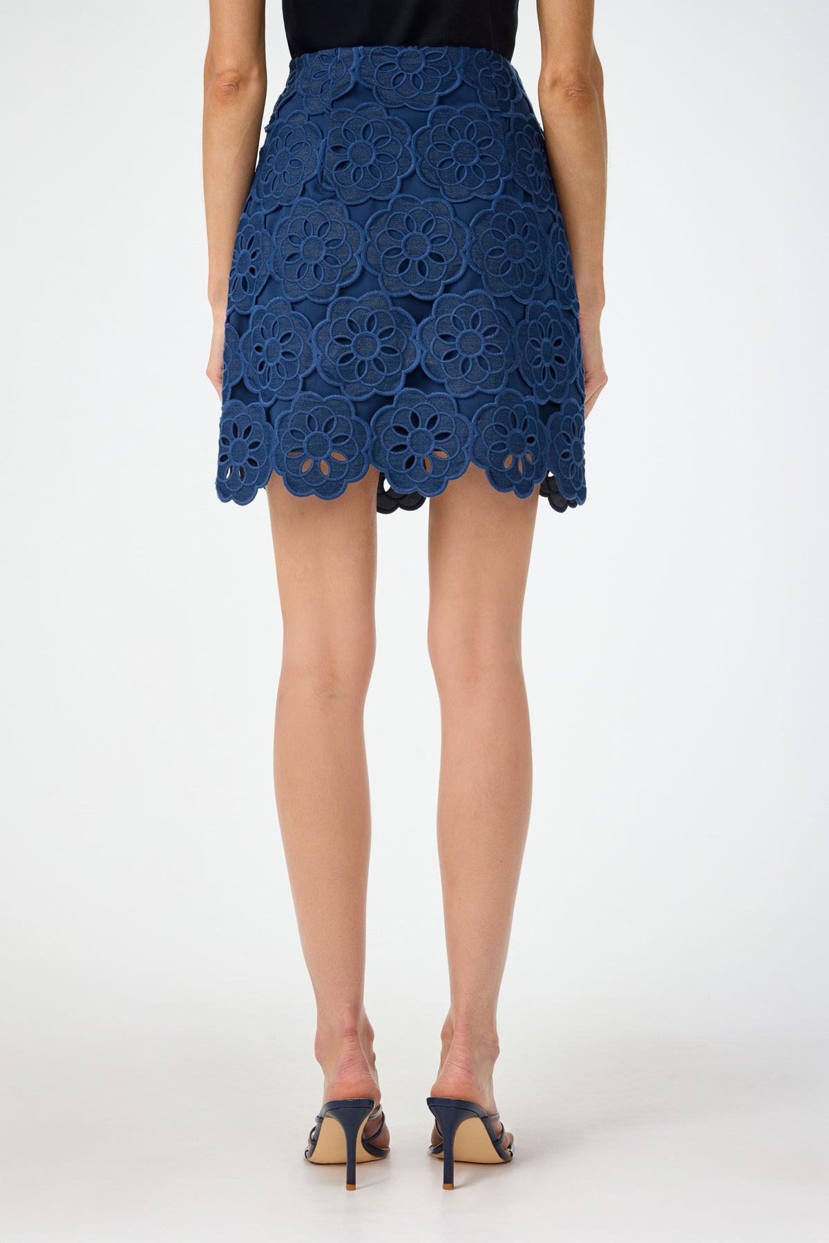 A person stands facing away, wearing the High Rise Rico Skirt - Kusho Embroidery with large floral lace patterns and a scalloped hem, paired with open-toe heeled sandals against a plain white background.