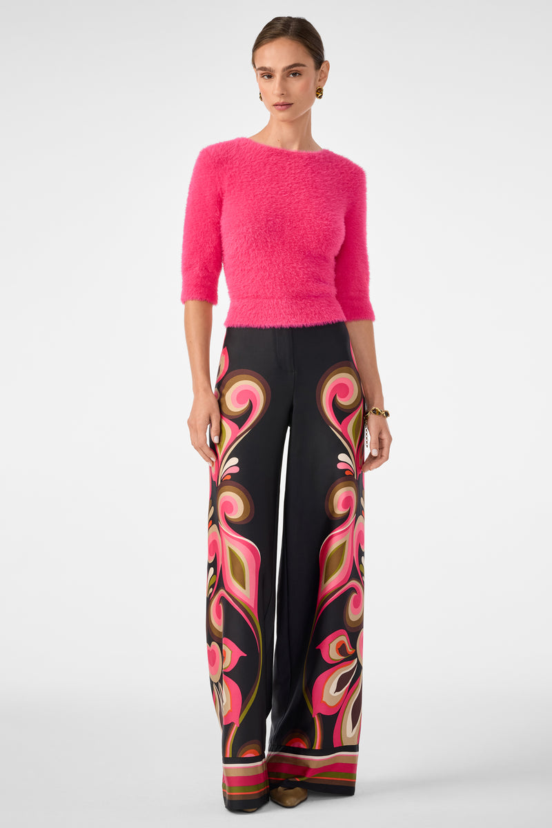 A woman wears the Maliah Fuzzy Sweater in bright pink and high-waisted, wide-leg black pants with a bold swirling pink, red, and beige pattern. Her hair is pulled back and she finishes the look with brown shoes.