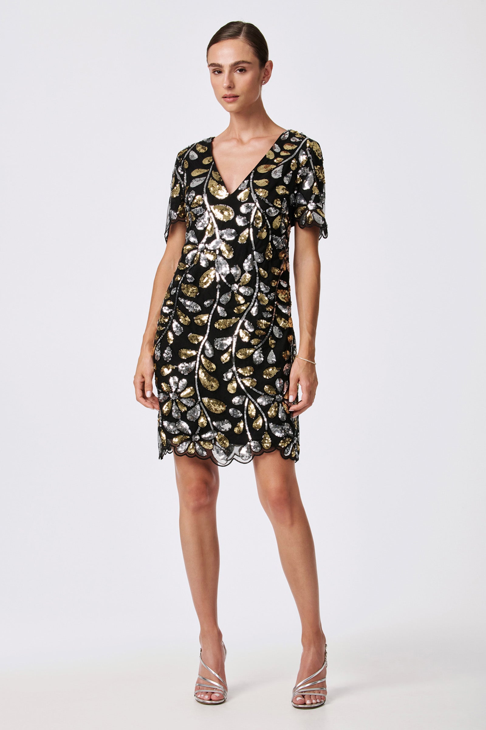 A woman poses against a plain background in the Obelle 2 Sequin Mini Dress, featuring black fabric with silver and gold leaf-shaped sequins, short sleeves, a V-neckline, and silver strappy heels.