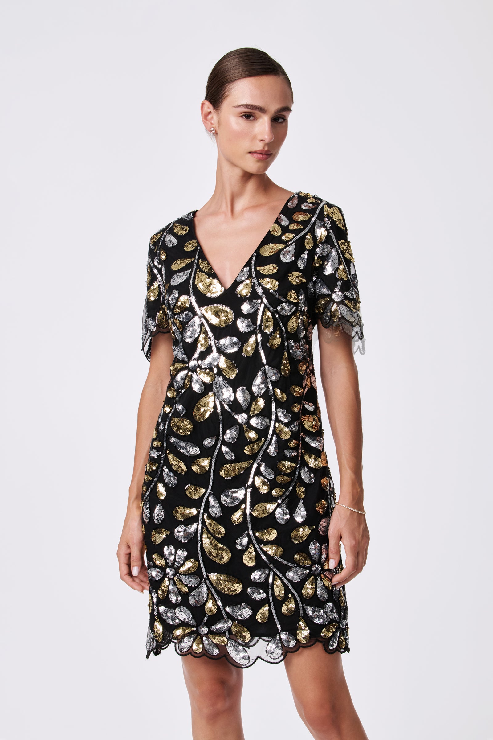 A woman stands against a plain light background wearing the OBELLE 2 DRESS, a black V-neck, short-sleeve dress made from 100% nylon with gold and silver sequin leaf patterns and a scalloped hem.