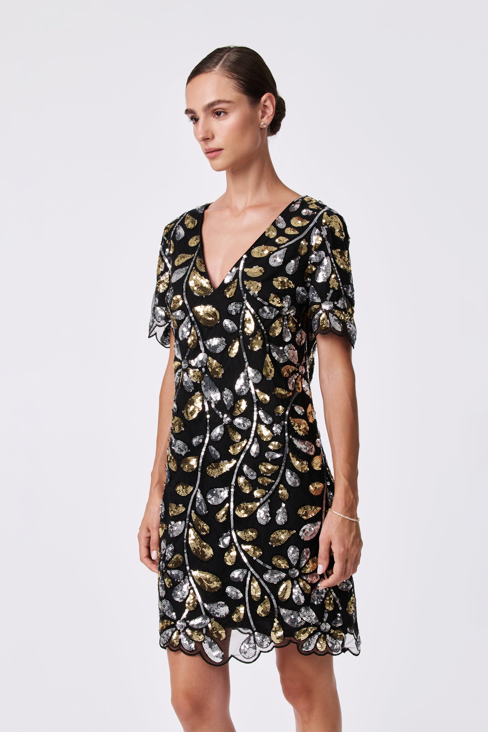 A woman stands against a plain background wearing the OBELLE 2 DRESS, a black 100% nylon dress with gold and silver sequin leaf patterns, short sleeves, and a V-neckline. Her hair is styled in a neat bun.