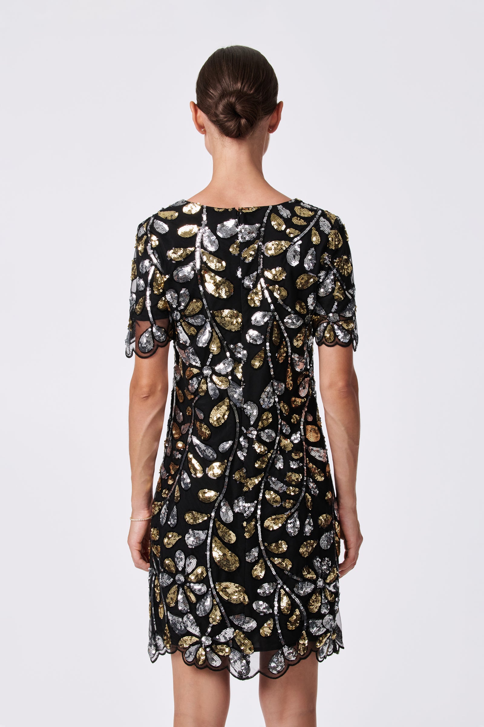 A woman with brown hair in a low bun models the OBELLE 2 DRESS, a short-sleeved black dress made of 100% nylon with gold and silver sequin leaf patterns, facing away against a plain white background.
