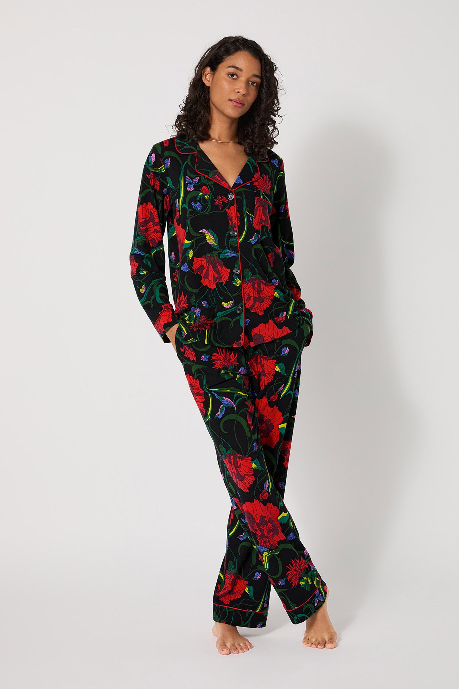 A woman stands barefoot against a plain background, wearing the Haute Floral L/S Classic Pj—matching black pajamas with a vibrant red, green, and blue floral print. Her hands are in her pockets as she looks slightly to the side.