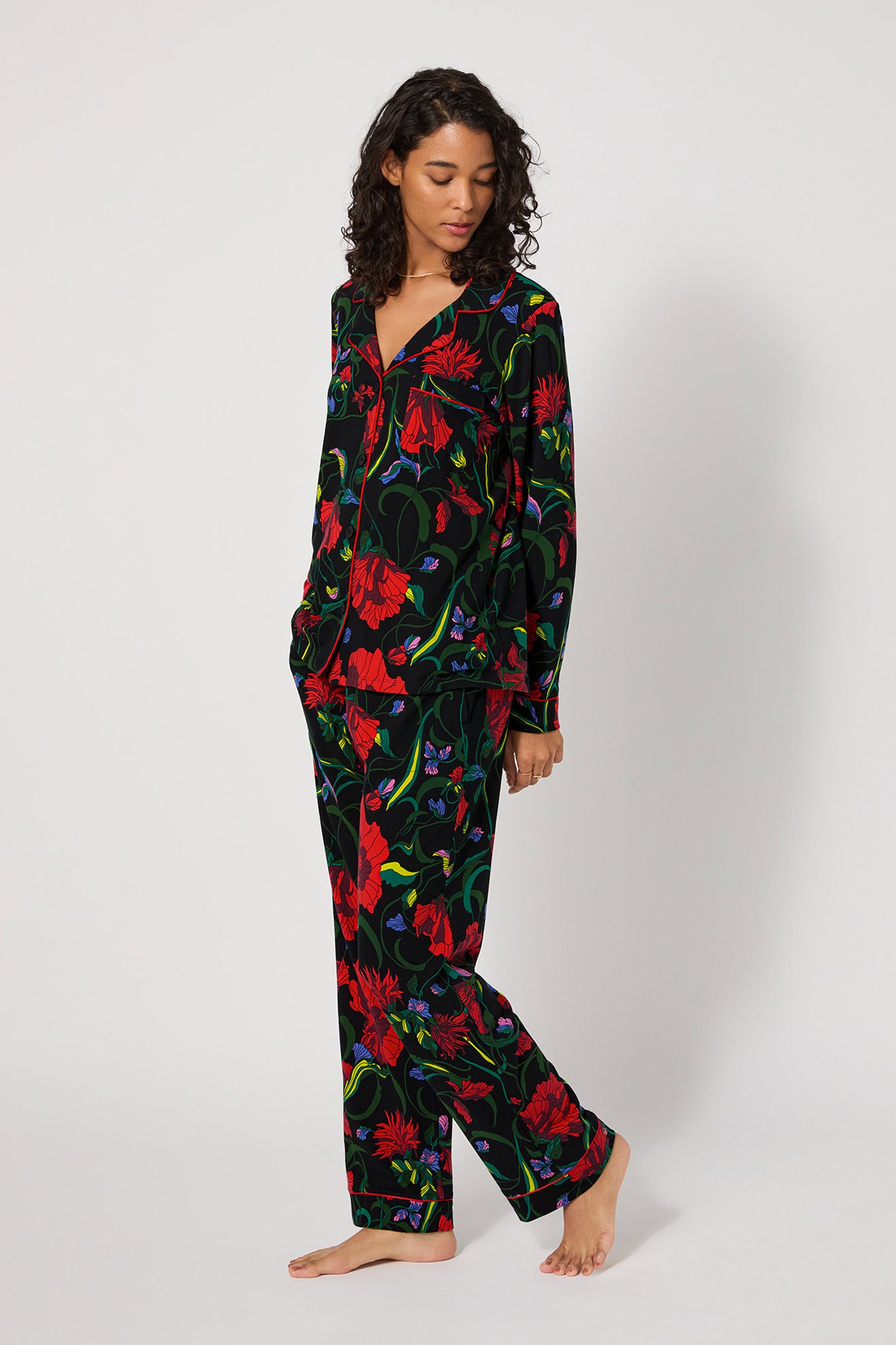 A woman with curly hair wears the Haute Floral L/S Classic Pj, a matching set with a vibrant red, purple, and green floral print on black. She stands barefoot against a plain white background, gazing down and to the side.