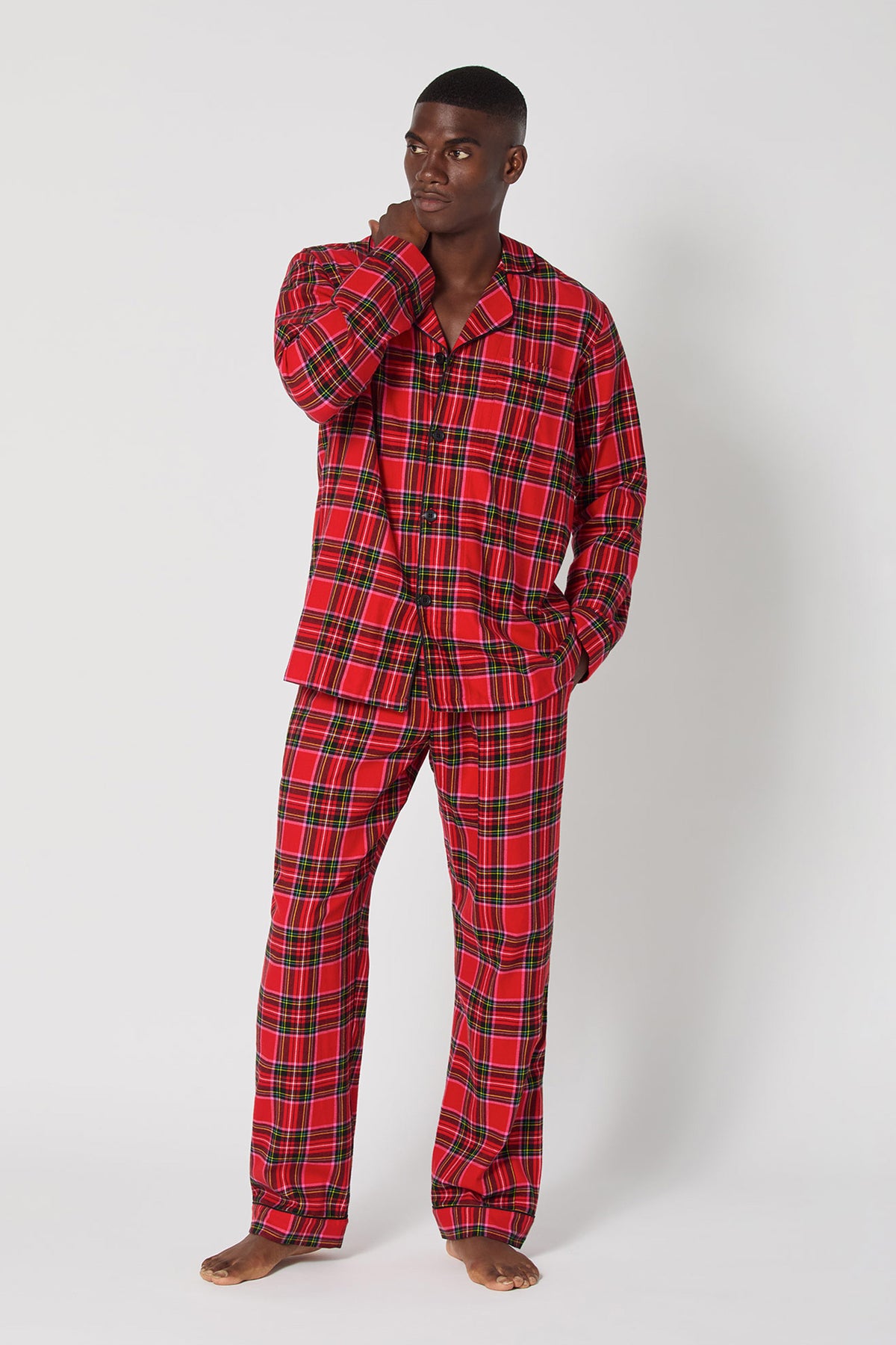 A man stands barefoot against a plain background, wearing the Christmas Plaid Men Classic Pj set with long sleeves and long pants. He looks to the side, touching his face with one hand.