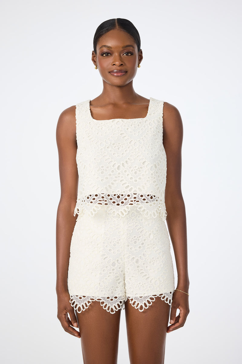 A woman wears the Albi Lace Crop Top in Whitewash, a sleeveless eyelet lace design, paired with matching high-waisted shorts, standing against a plain white background.