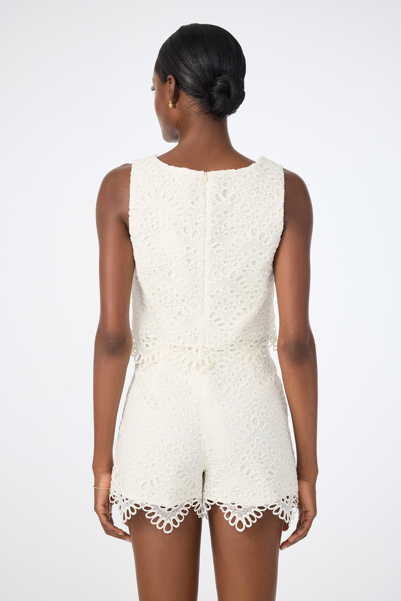 A woman with dark hair in a low bun is seen from behind, wearing the Albi Lace Crop Top—a sleeveless, white lace top with scalloped edges and intricate cut-out patterns—against a plain light background.