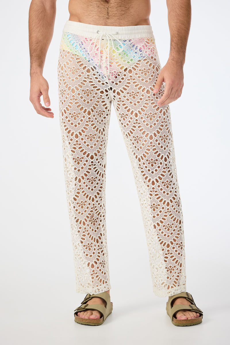 A person wears the Venice Beach Resort Pant - Lerins Lace in Whitewash, featuring a see-through crocheted lace design and drawstring waist. Styled with brown sandals, the detailed pattern hints at colorful underwear underneath.
