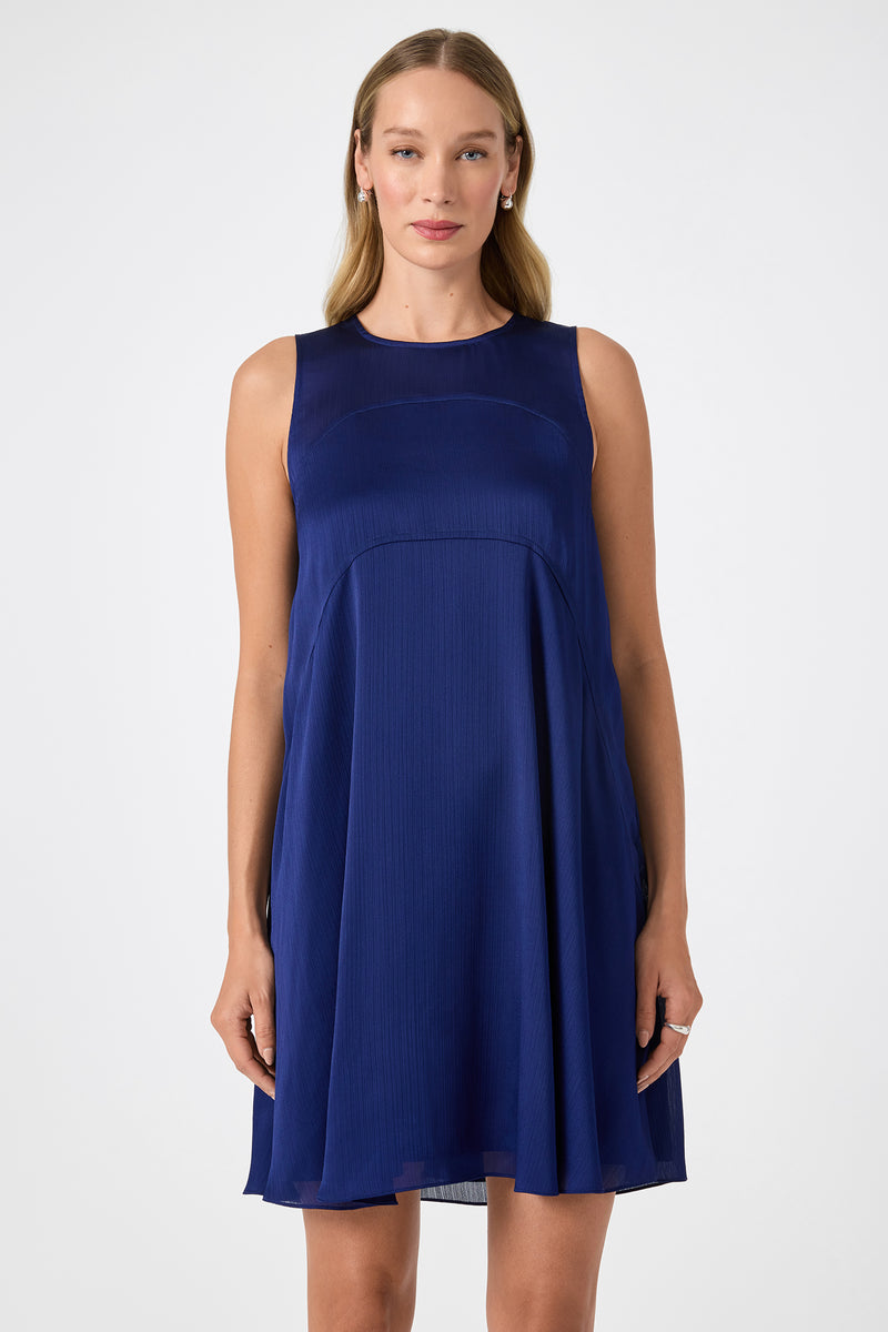 A woman with long blonde hair wears the Mauvie Sleeveless Mini Dress, a dark blue knee-length piece. She stands facing forward against a plain white background with a neutral expression.