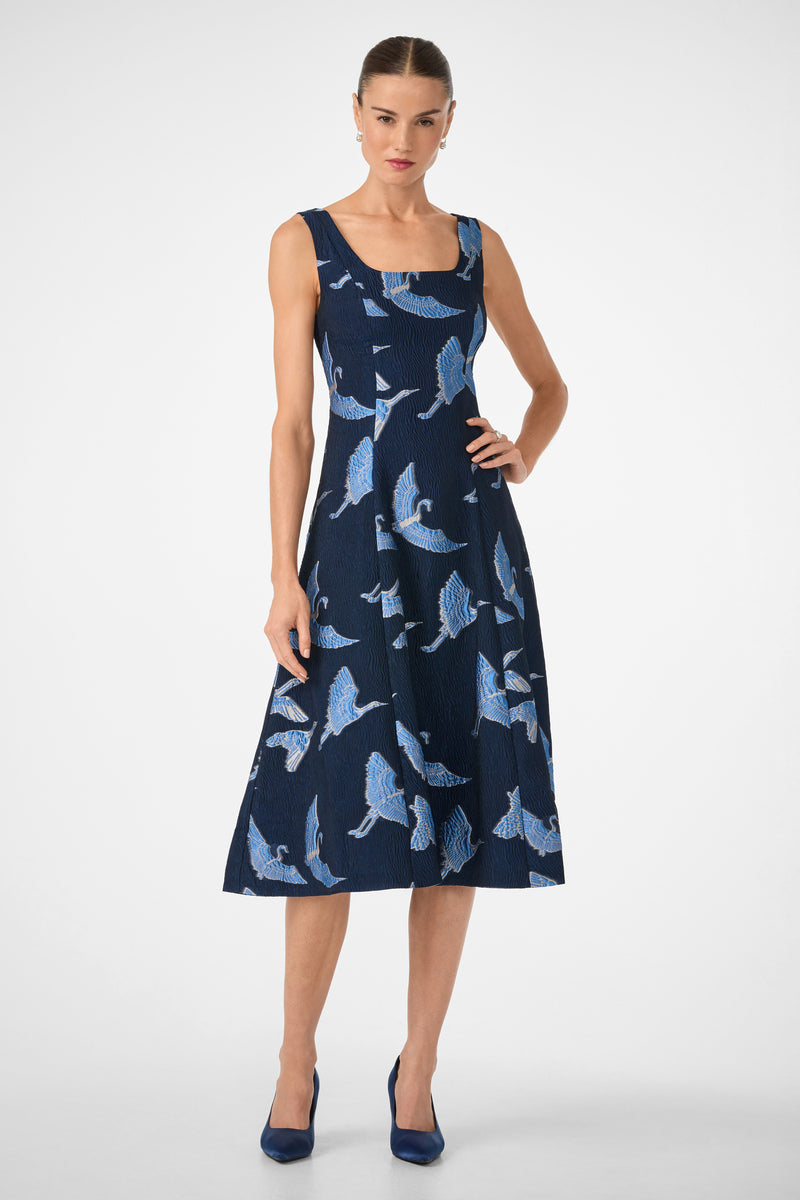 A woman wears the Valor Sleeveless Midi Dress, a navy blue midi with a square neckline, flared skirt, and blue-and-white bird print. She stands against a white background and pairs it with matching blue heels.