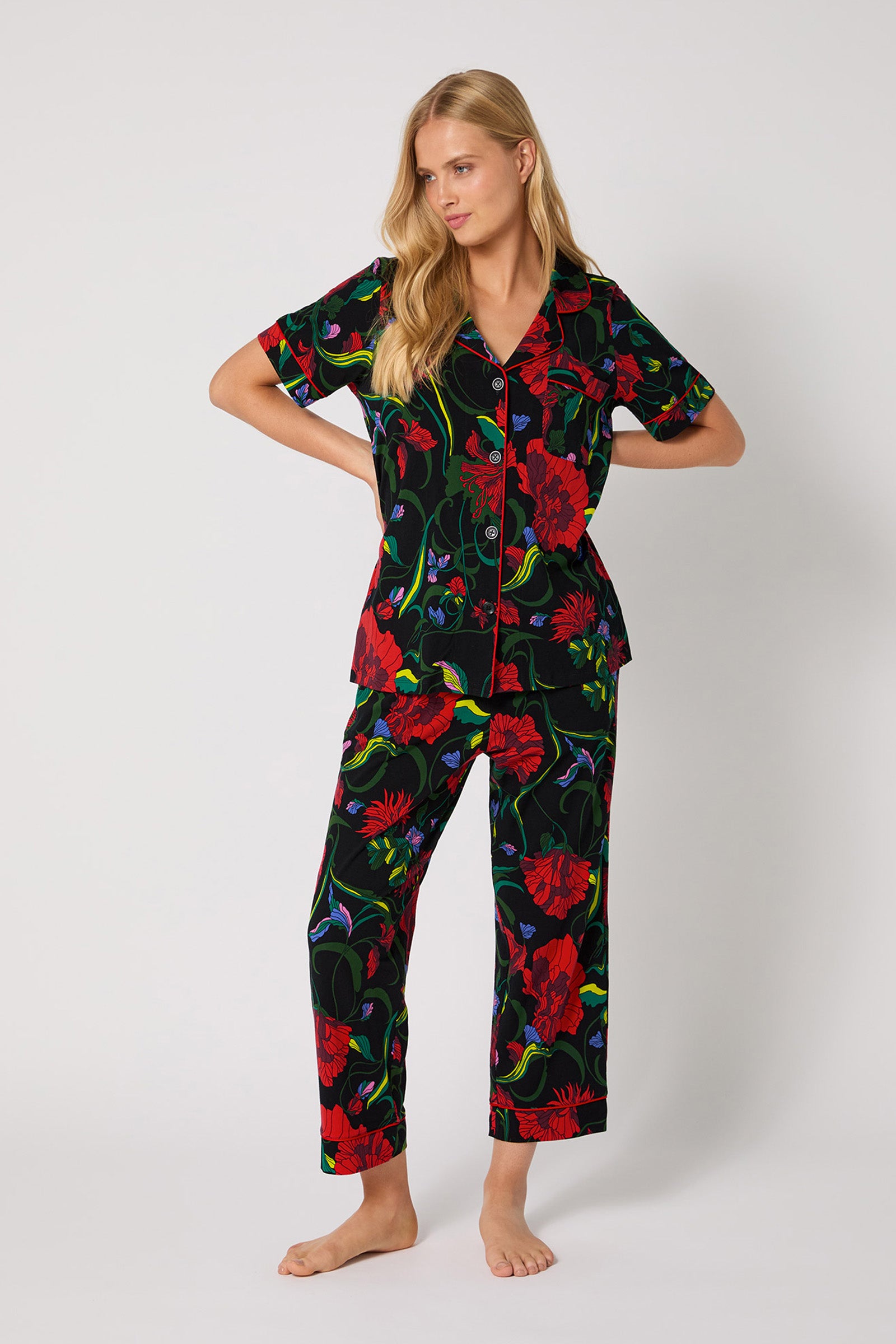 A woman with long blonde hair models the Haute Floral S/S Cropped PJ Set, featuring a black background and vibrant red floral print. She stands barefoot against a plain light backdrop, one hand on her hip.
