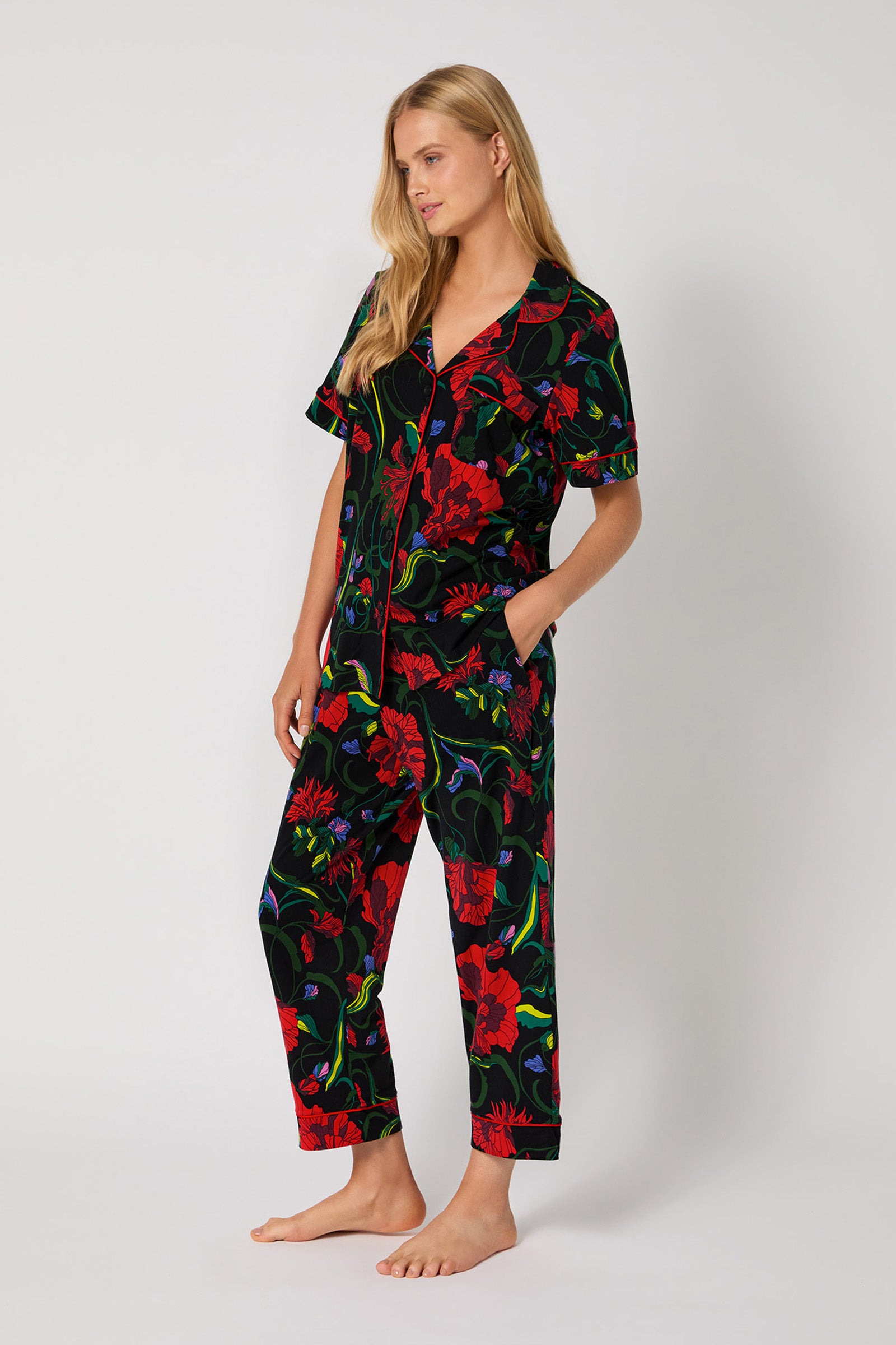 A woman stands barefoot against a plain background, wearing the Haute Floral S/S Cropped Pj Set—a matching short-sleeved, button-up pajama set featuring a bold red, green, and purple floral print on black. One hand rests in her pocket.