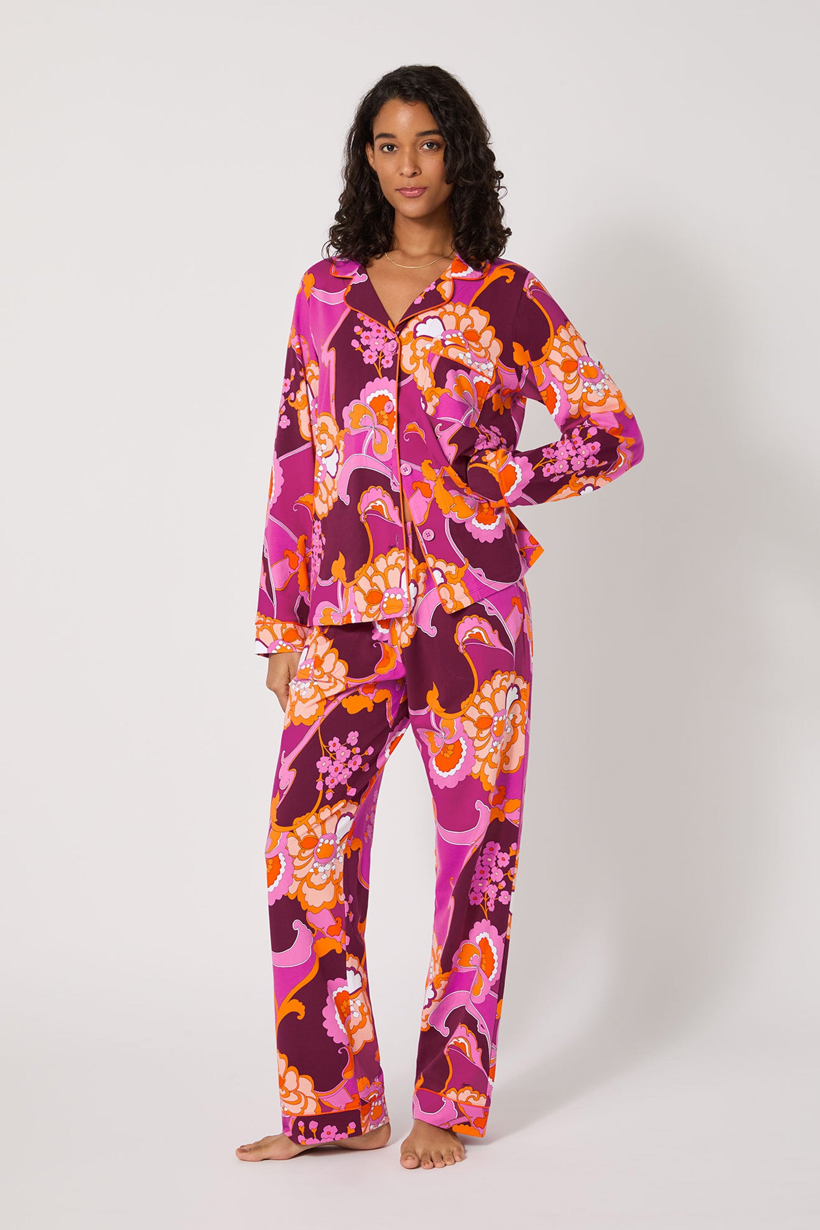 A woman stands barefoot against a plain background, wearing the Apache Plume L/S Classic Pj—a vibrant pajama set featuring a bold floral pattern in shades of pink, orange, purple, and white.