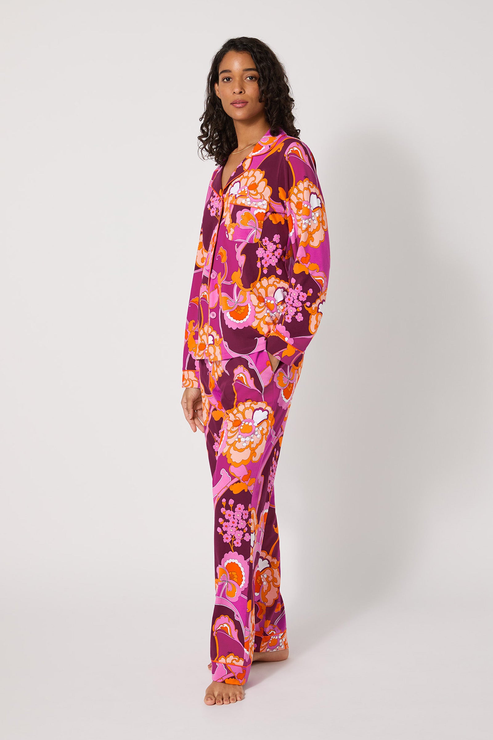 A woman with curly hair stands barefoot against a white background, wearing the Apache Plume L/S Classic Pj—a matching set with a vibrant floral pattern in pink, orange, red, and white—and looks relaxed.
