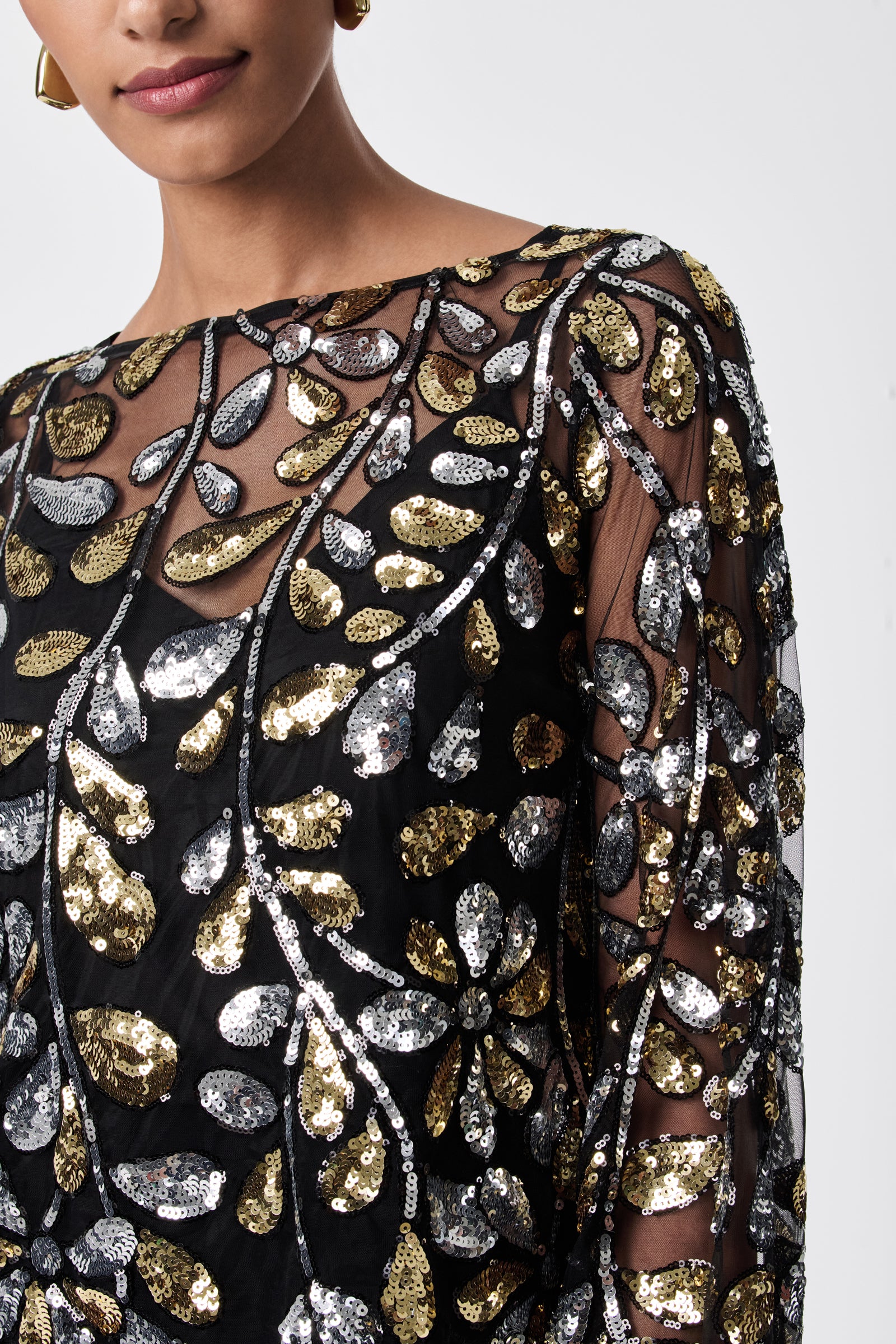 A close-up of a person wearing the FEMMES TOP—a sheer black 100% nylon top with gold and silver sequin leaf patterns, long sleeves, and a round neckline. Only the lower half of the face and upper torso are visible.